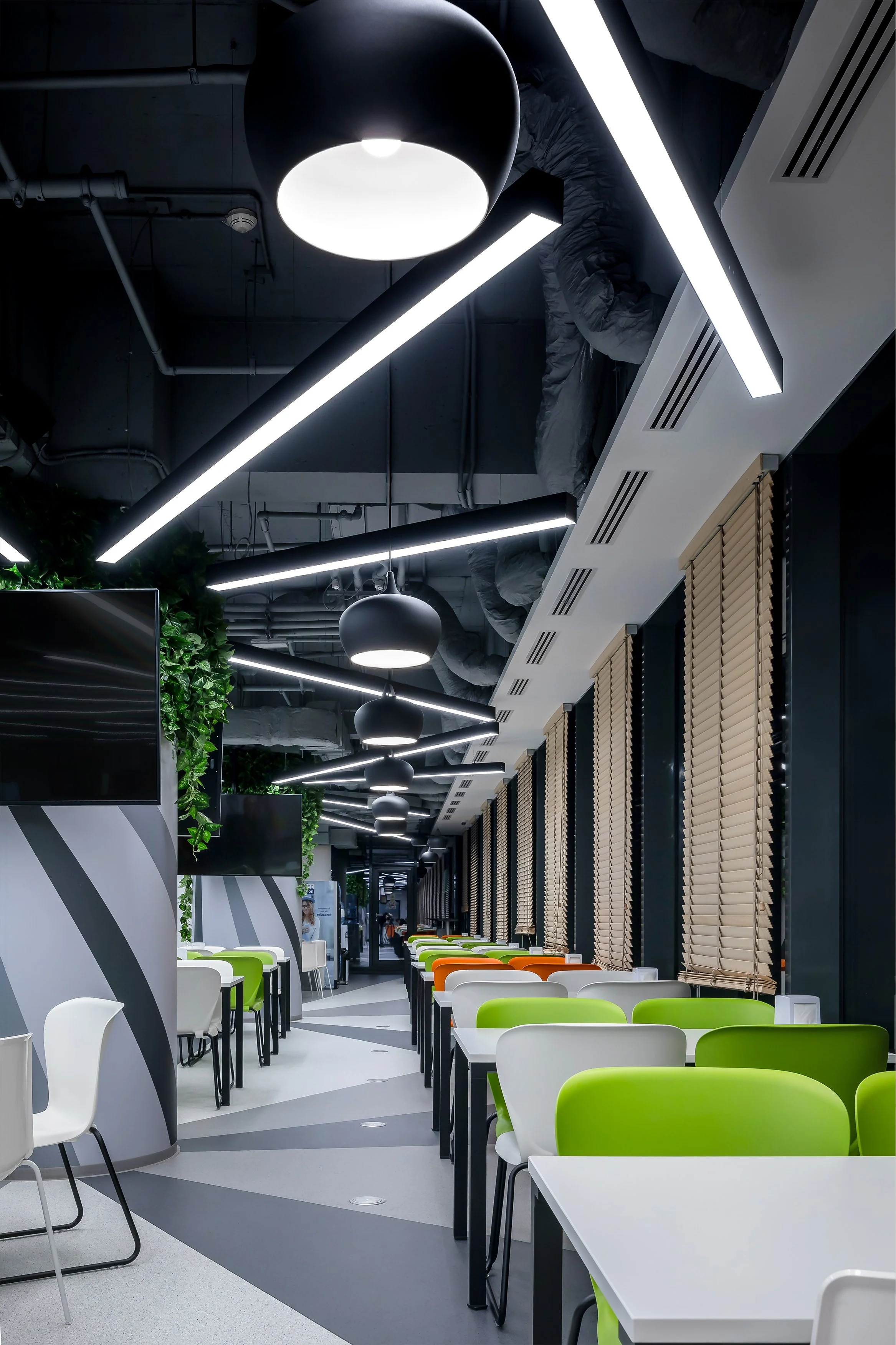 Modern cafeteria with white, green, and orange chairs, rectangular tables, and decorative blinds on windows. Ceiling has black pendant lamps and white linear lights, and walls feature greenery and TV screens.