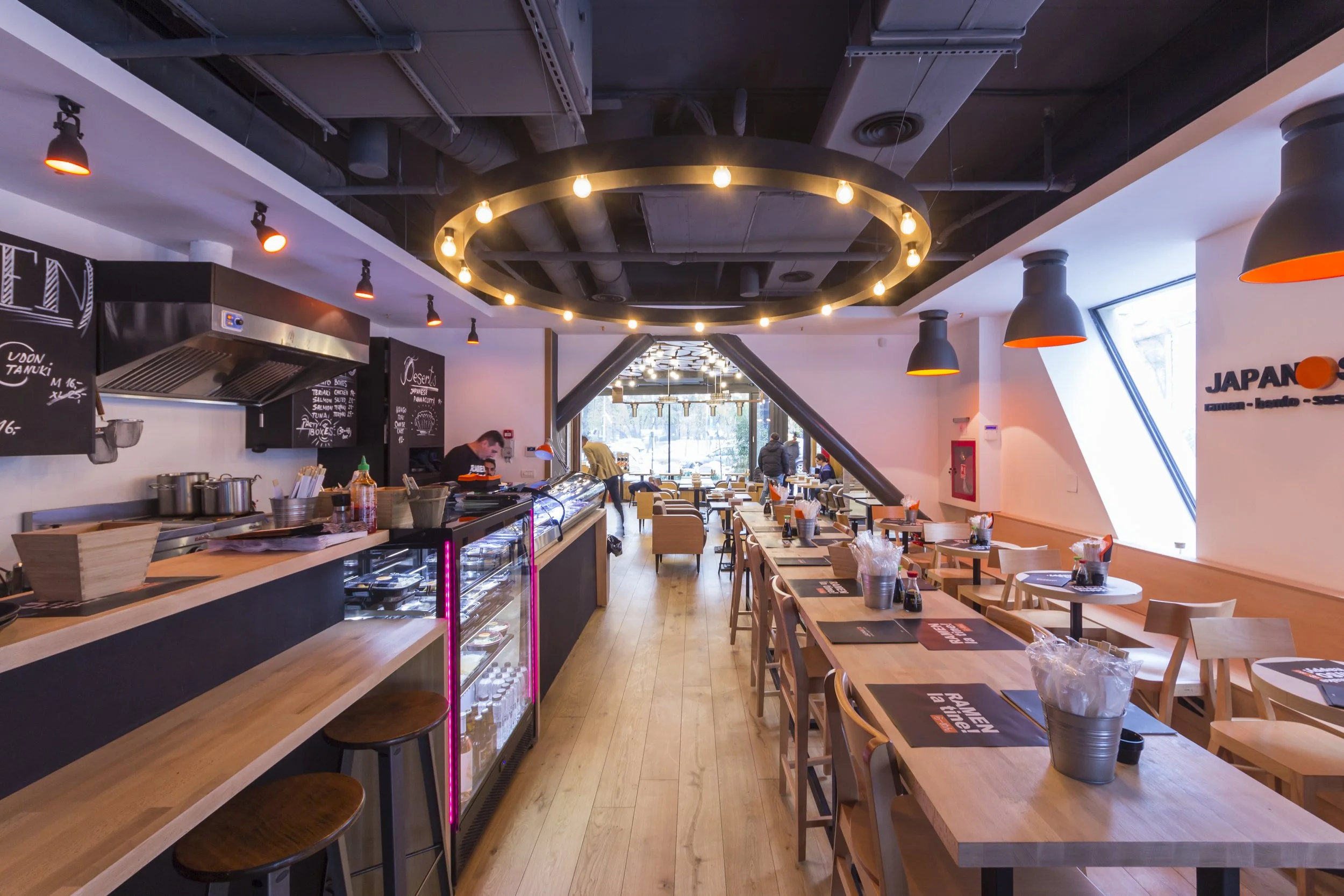 Interior of a modern restaurant with wooden tables, chairs, and black and orange lighting fixtures, featuring a bar area on the left and large windows on the right letting in natural light.
