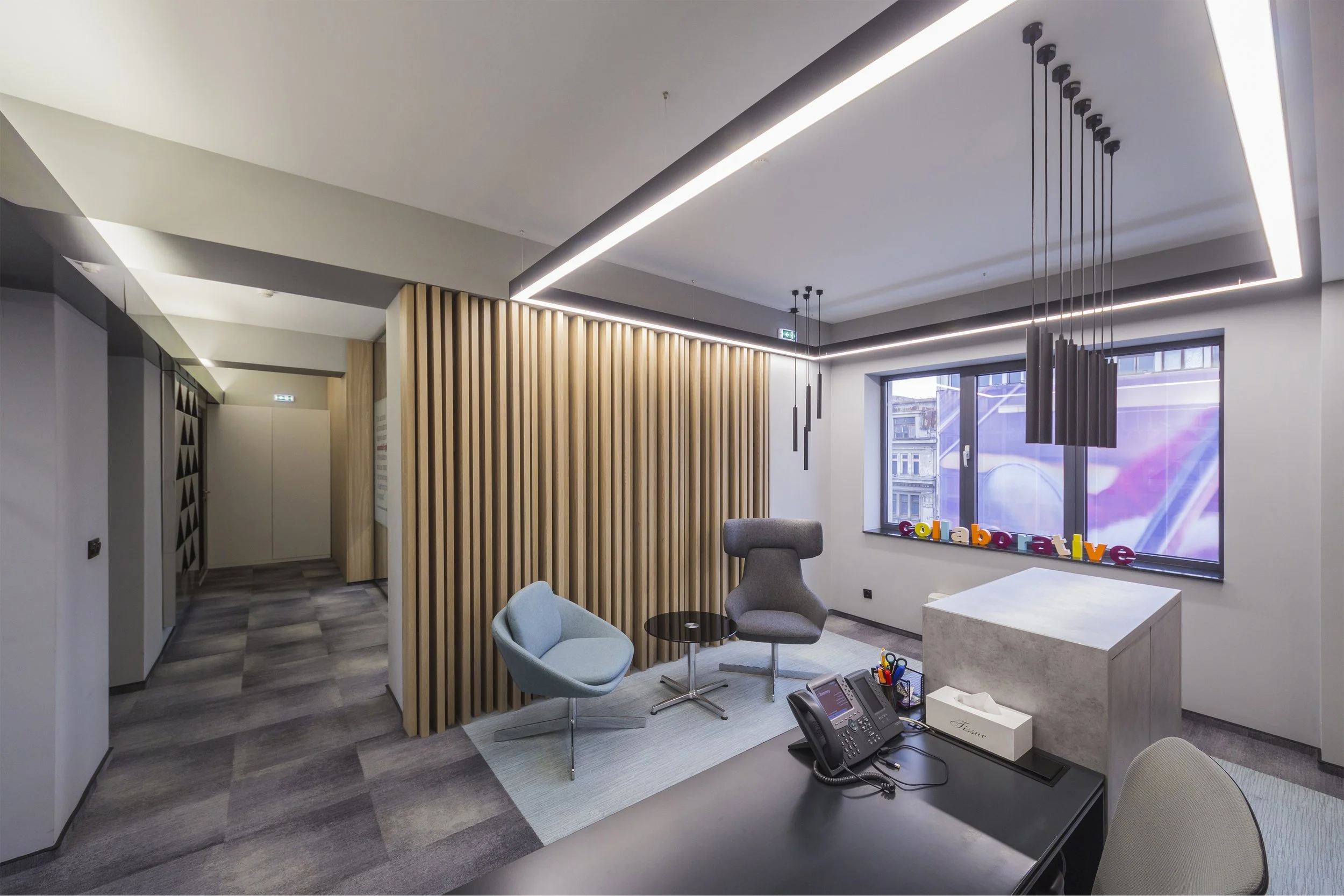 Modern office reception area with seating, a desk phone, colorful 'collaborative' letters on the windowsill, and contemporary lighting fixtures.