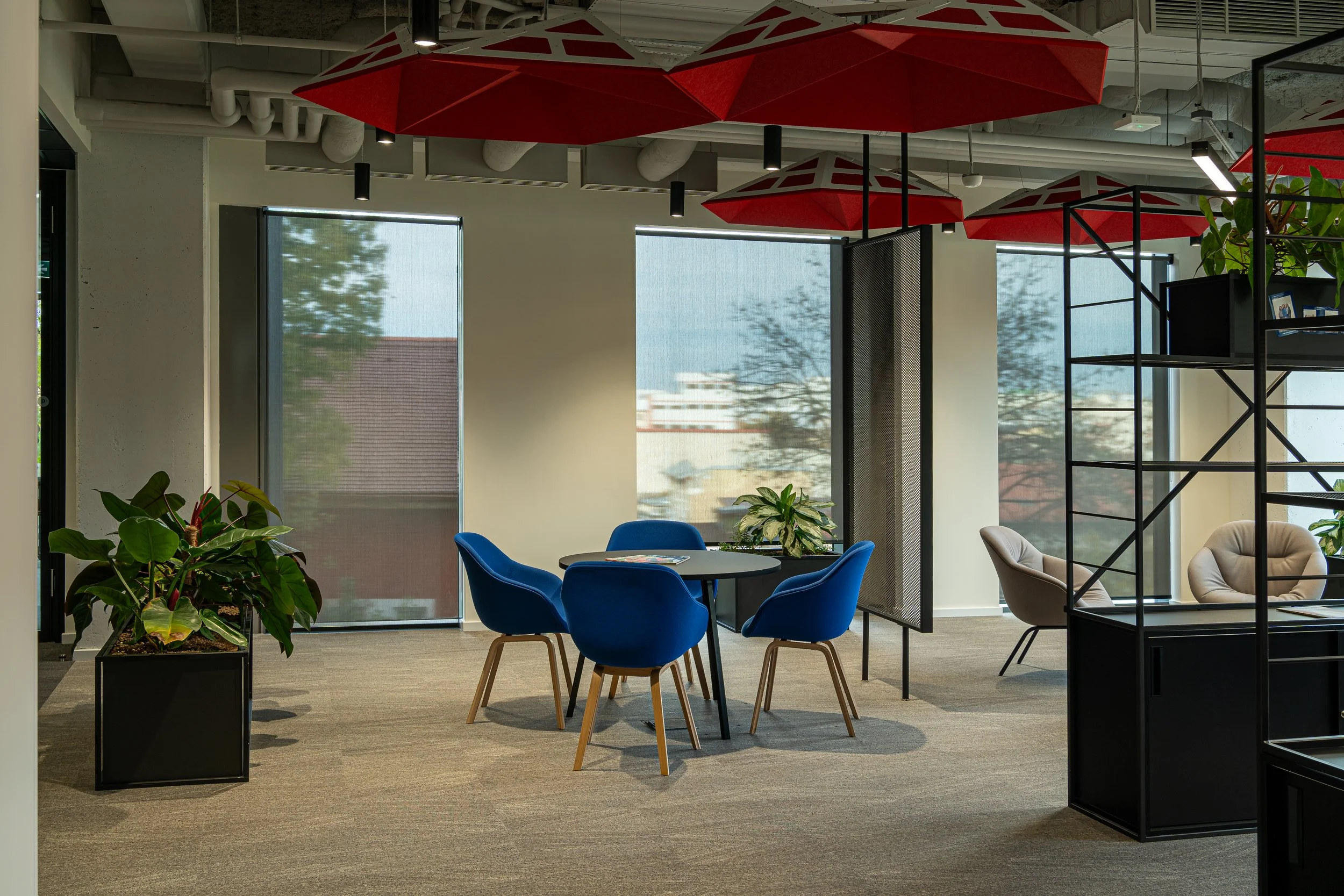 Modern office interior with a round table surrounded by four blue chairs, two beige lounge chairs, large windows, potted plants, and red umbrellas on the ceiling.