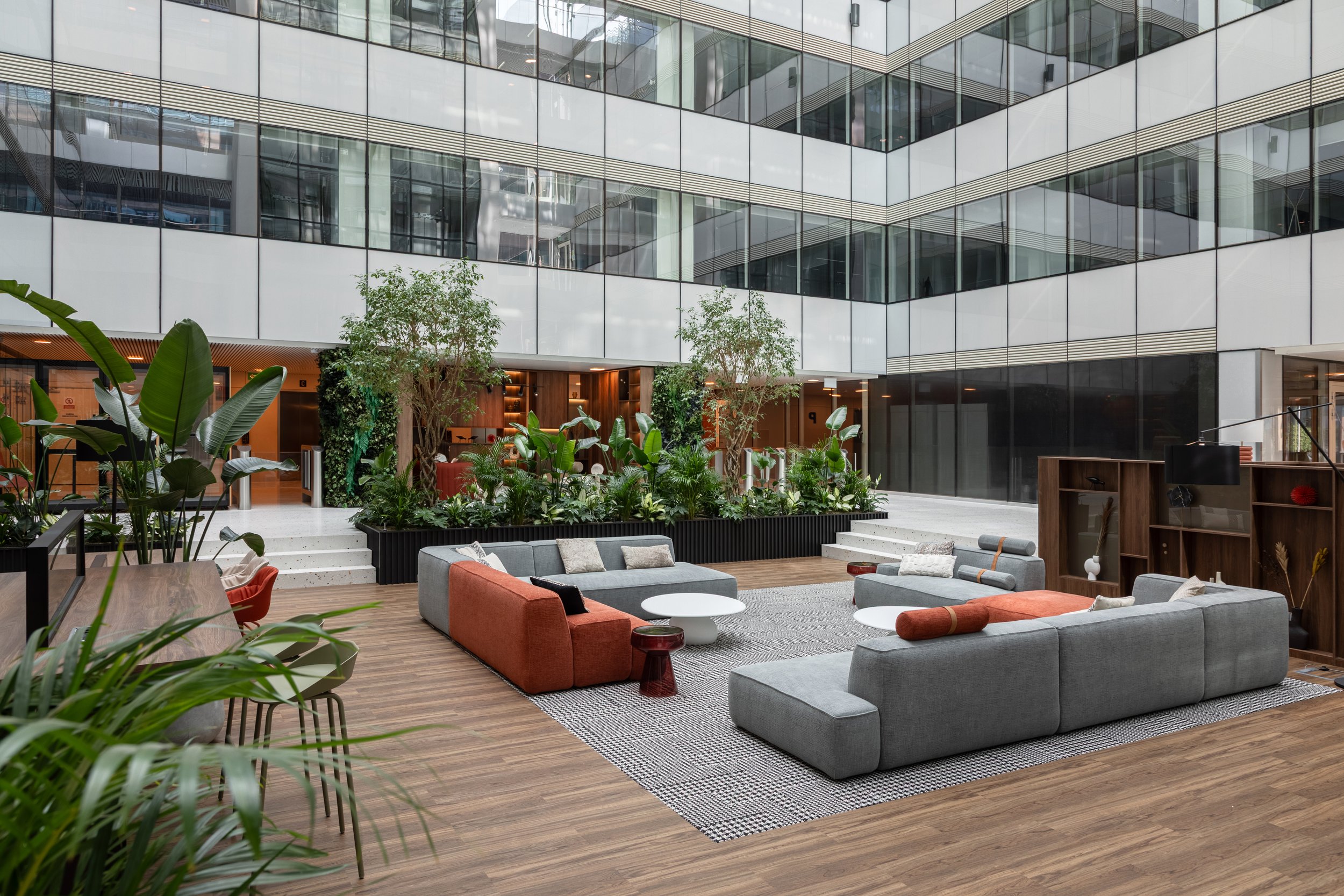 Modern hotel lobby with high glass ceilings, green plants, and contemporary furniture including gray and orange sofas and small tables on wooden flooring.