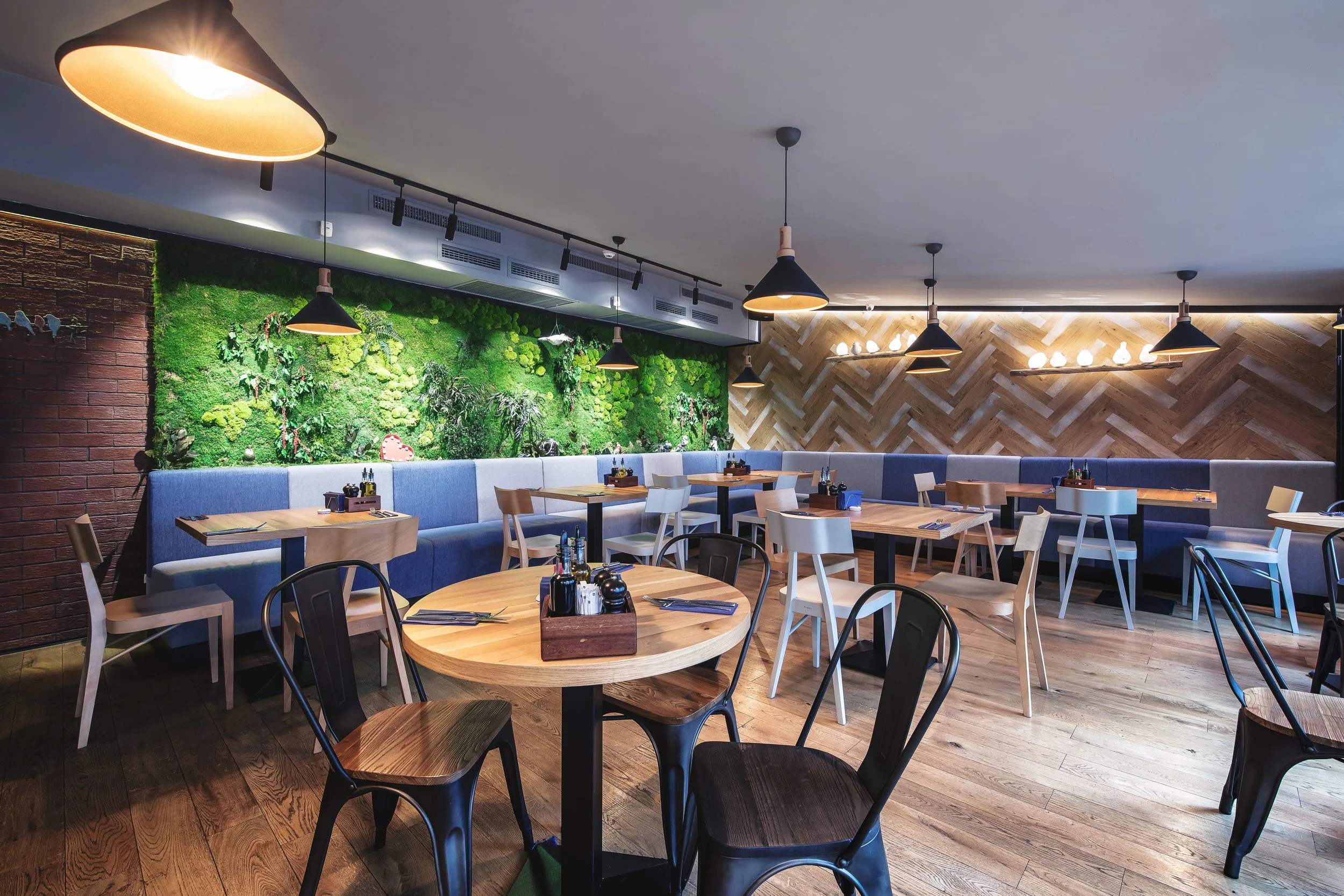 Modern restaurant interior with wooden floors, a green moss wall, and a mix of black, white, and natural wood chairs surrounding tables, with pendant lights overhead.