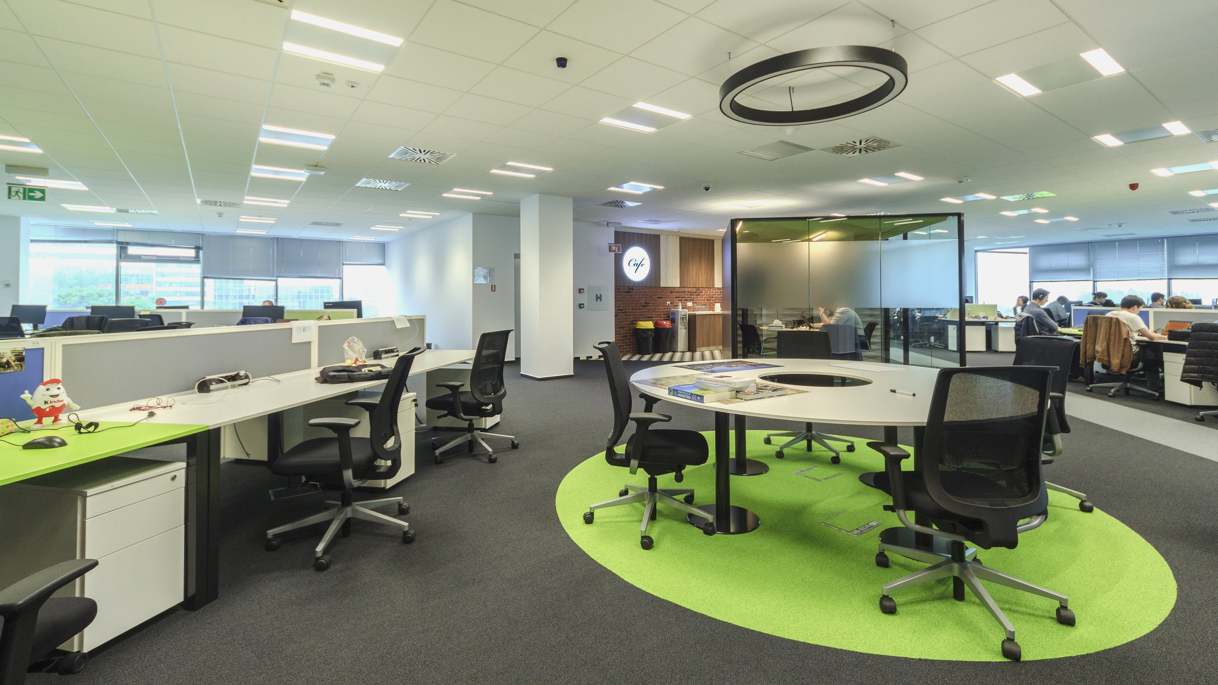 Open office space with desks, chairs, and large windows. A round table with chairs is in the foreground. In the background, employees work at computer desks, and a glass-walled meeting room is visible.