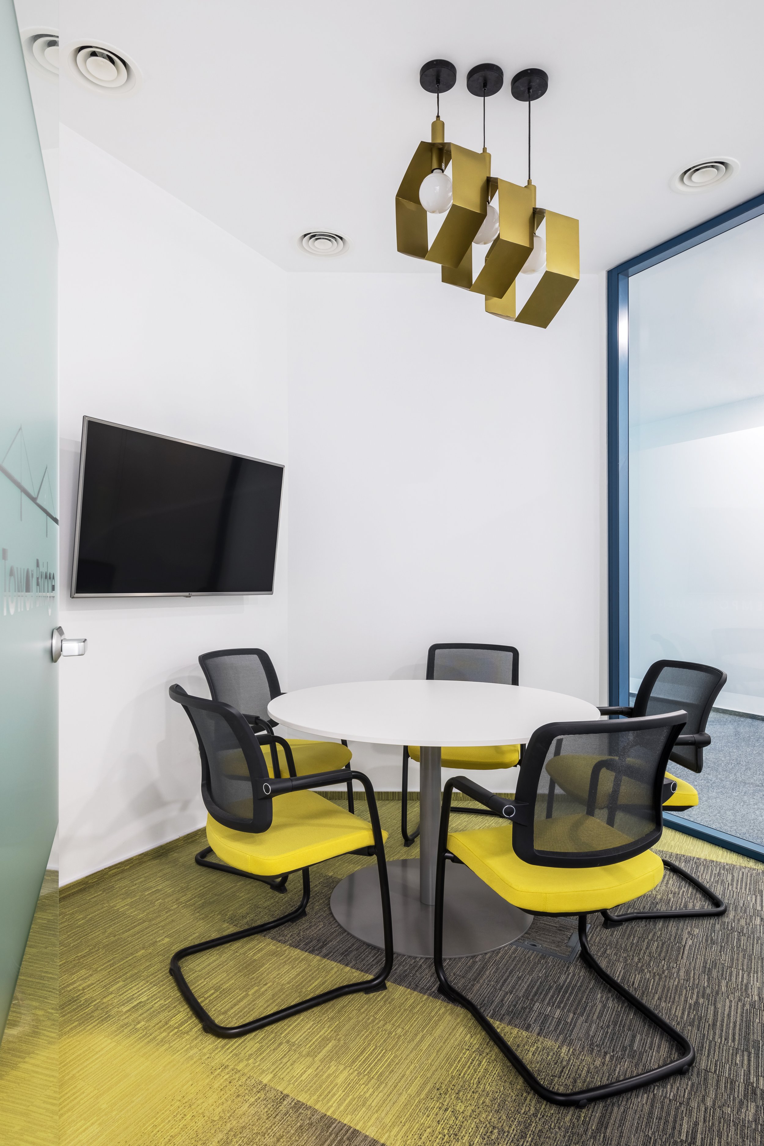 A modern conference room with a round white table, four black and yellow chairs with mesh backs, a wall-mounted flat-screen TV, and a gold geometric pendant light fixture on the ceiling. There's a large window with blue trim and a green and gray patterned carpet.