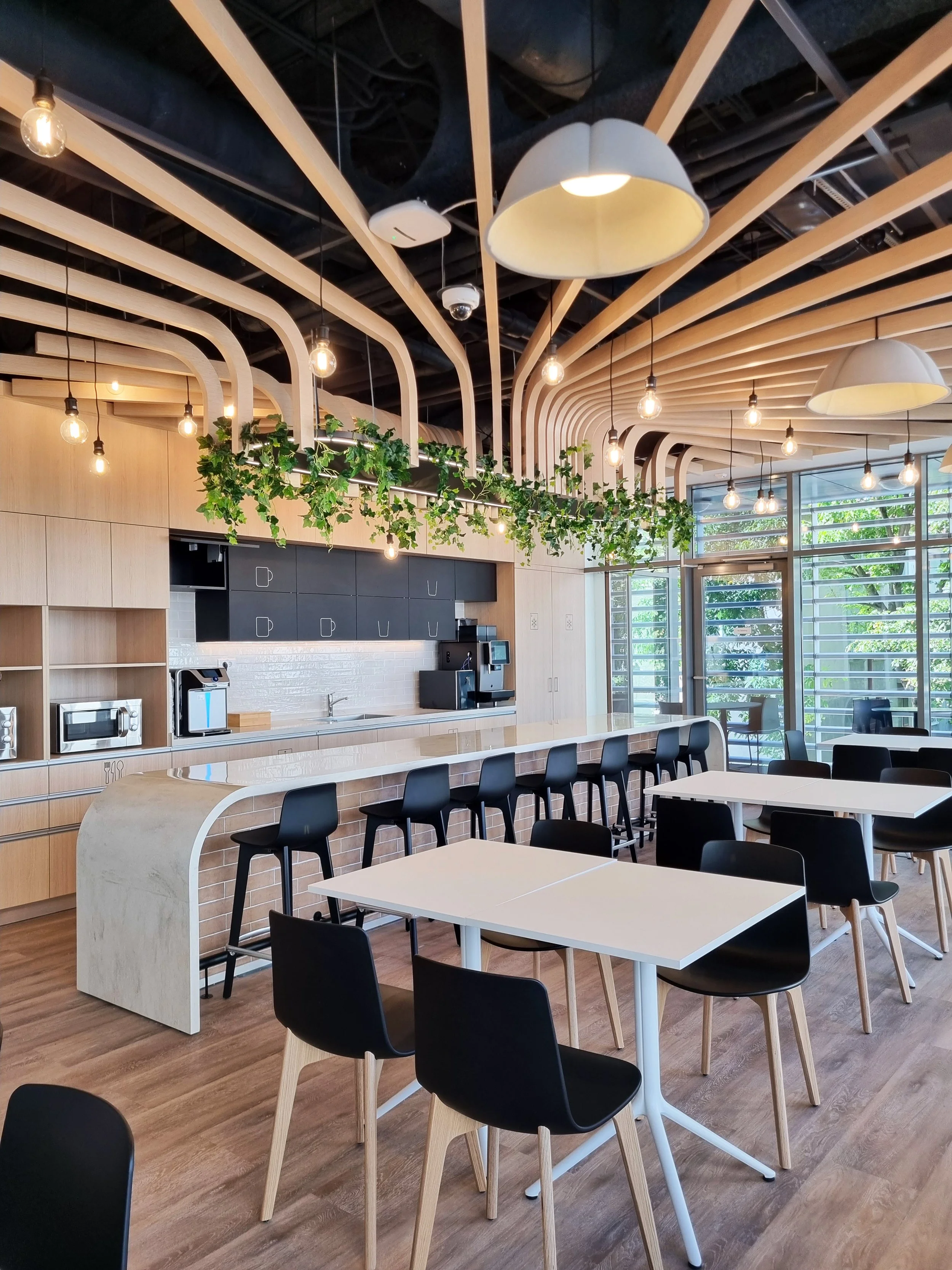 Modern cafe interior with white tables, black chairs, a bar area with black and white cabinets, wood accents, hanging light bulbs, and large windows with greenery outside.