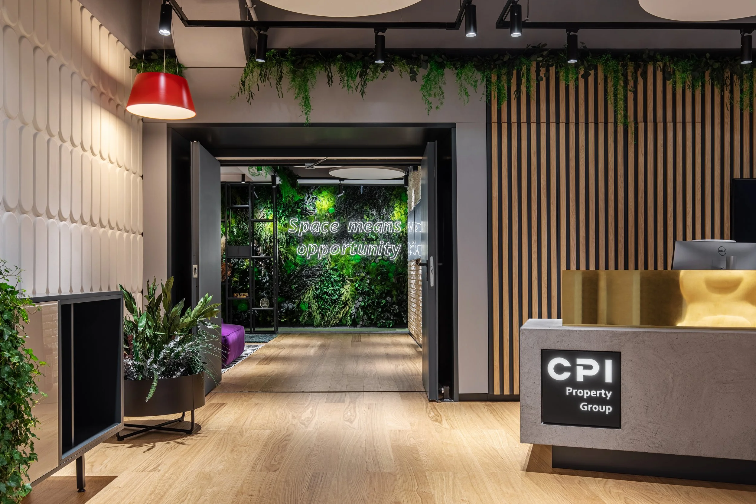 Modern office reception area with a white stone desk labeled 'CPI Property Group', wooden wall accents, green plants, a red pendant light, and a neon sign that reads 'Space means opportunity' in a green, plant-filled room beyond.