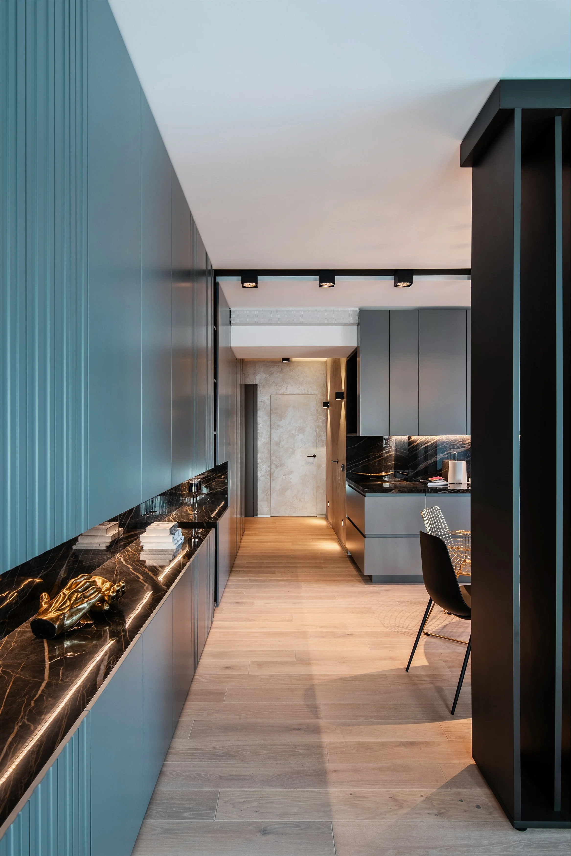 Modern kitchen with black marble countertops, blue and gray cabinetry, and wooden flooring. Small decorative items on the counter and a black chair near a black shelving unit.