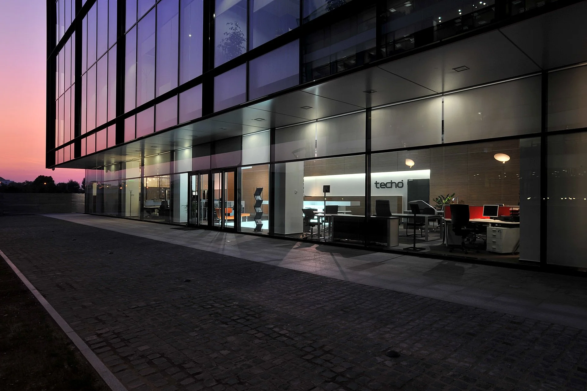 Modern office building with glass walls illuminated from inside during twilight, with visible office furniture and the sign 'techo' inside.