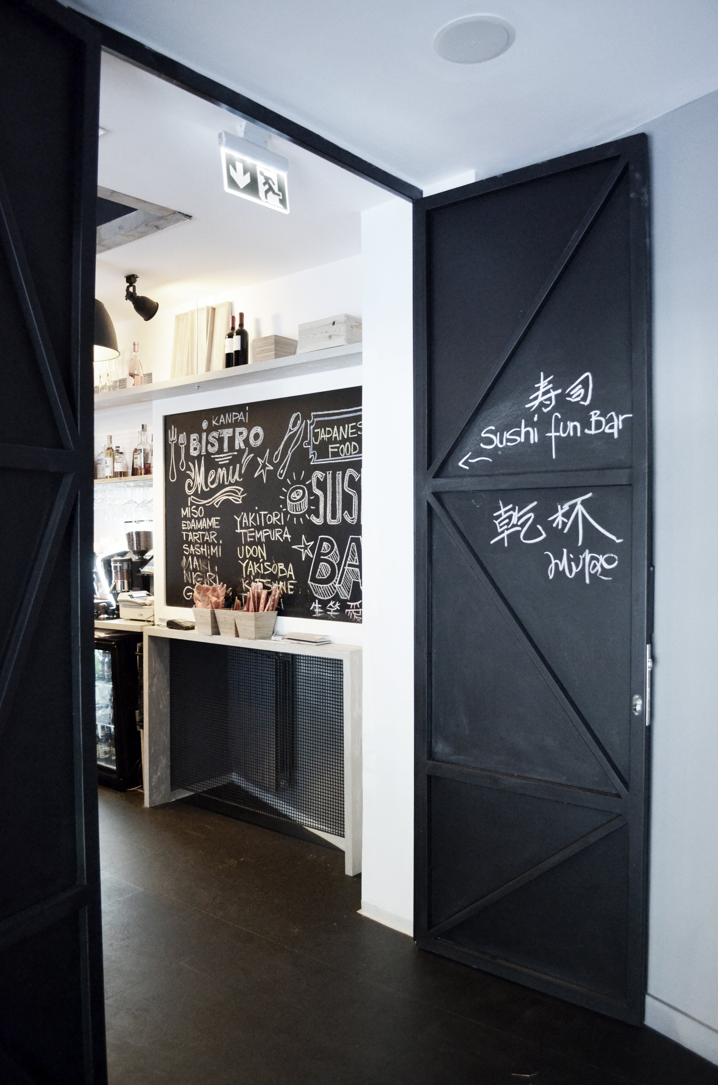 Interior view of a Japanese restaurant or bar, featuring a black chalkboard menu with various Japanese food items and a black door with white handwritten signs indicating sushi fun bar and a drink station.