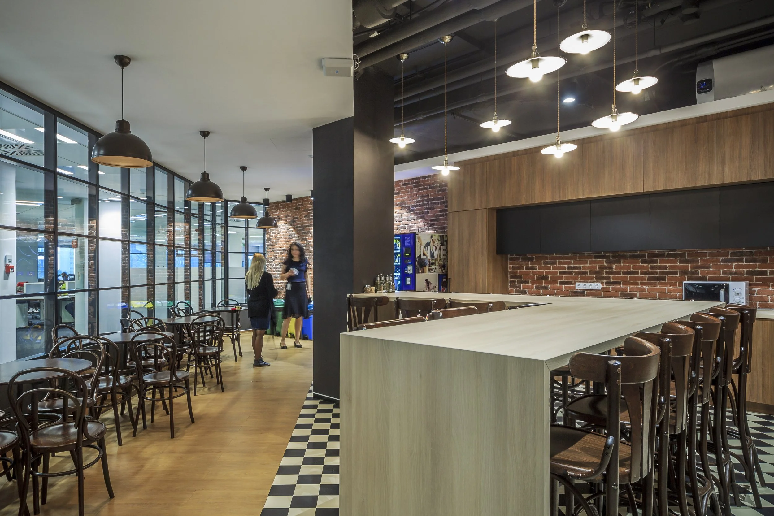 Interior of a modern café with wooden and brick walls, black ceiling, hanging lamps, and a checkered tile floor next to a wooden counter. There are patio-style chairs and tables, and two women standing near a snack machine at the back.