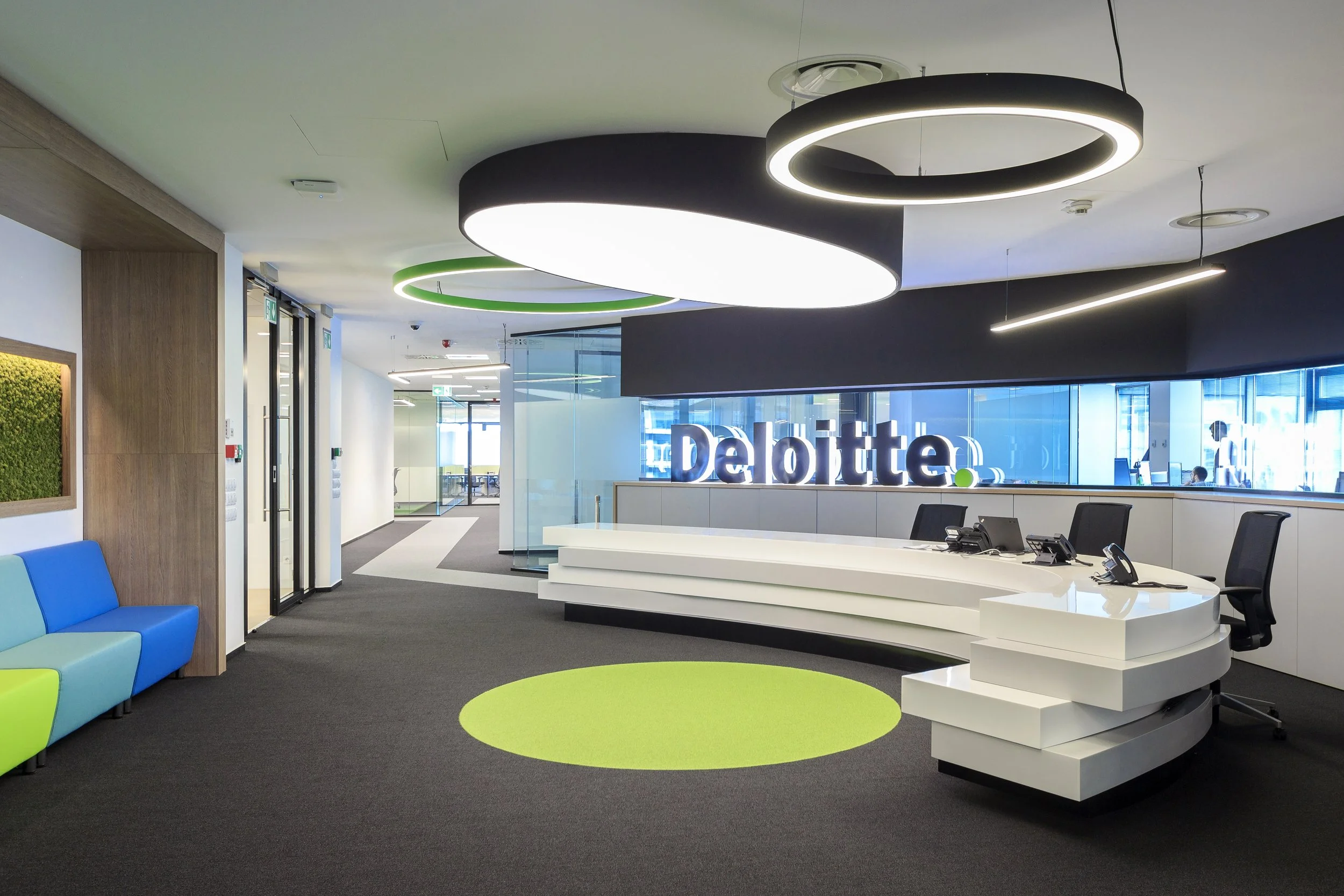 Modern office reception area with a curved white desk, black chairs, a Deere sign with illuminated letters, colorful ceiling lights, a green circular rug, and a glass wall.