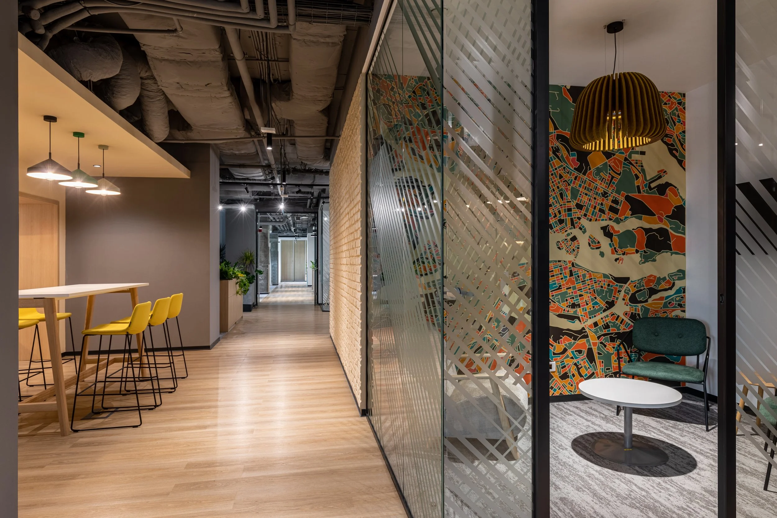 Modern office corridor with wooden flooring, glass partitions, colorful mural on wall, and a cozy seating area with green chair, white table, hanging yellow lamp, and decorative wall art.