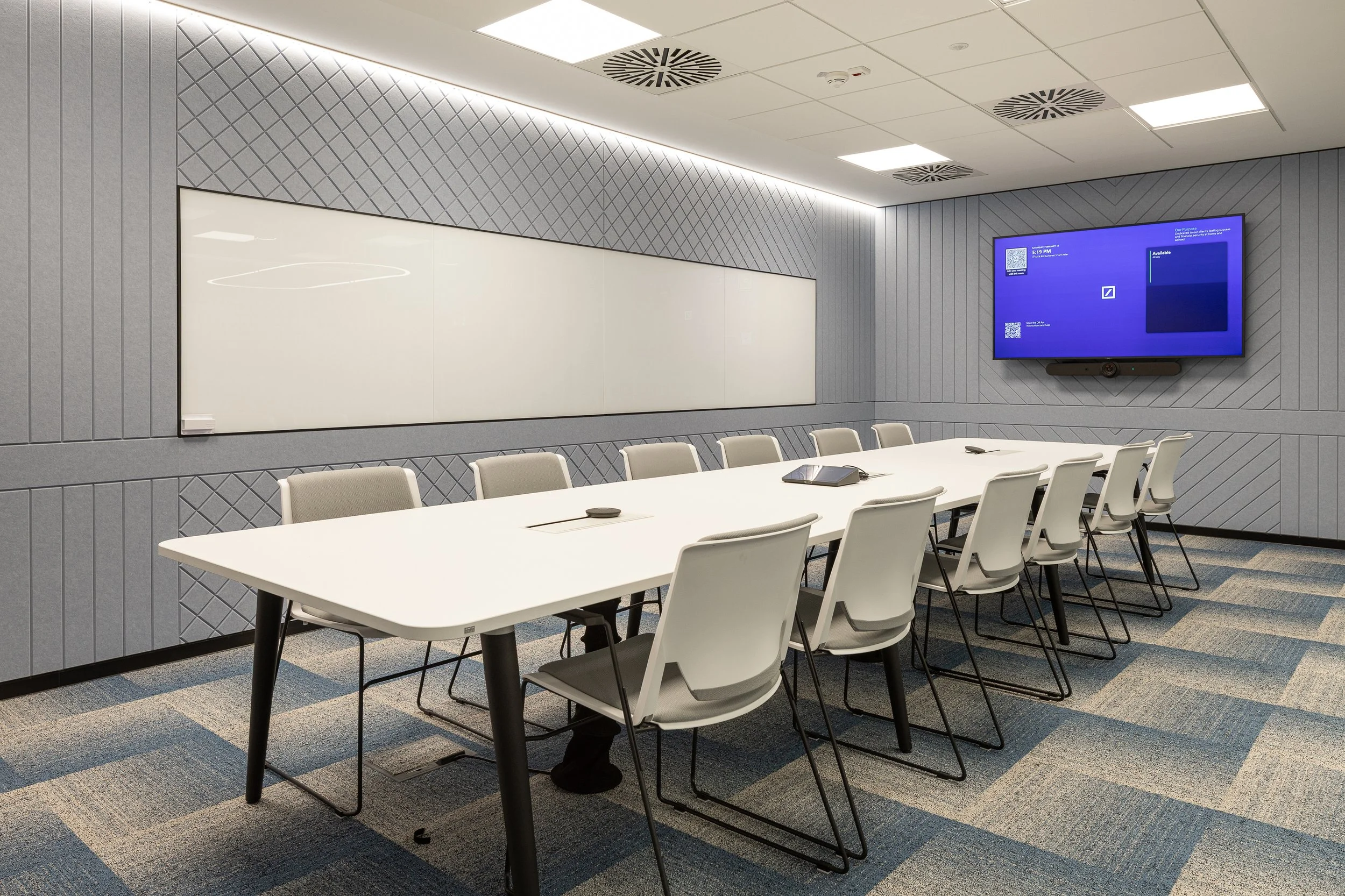 Empty conference room with a long white table surrounded by white chairs, a large whiteboard on the wall, and a flat-screen TV on the wall displaying a blue screen.