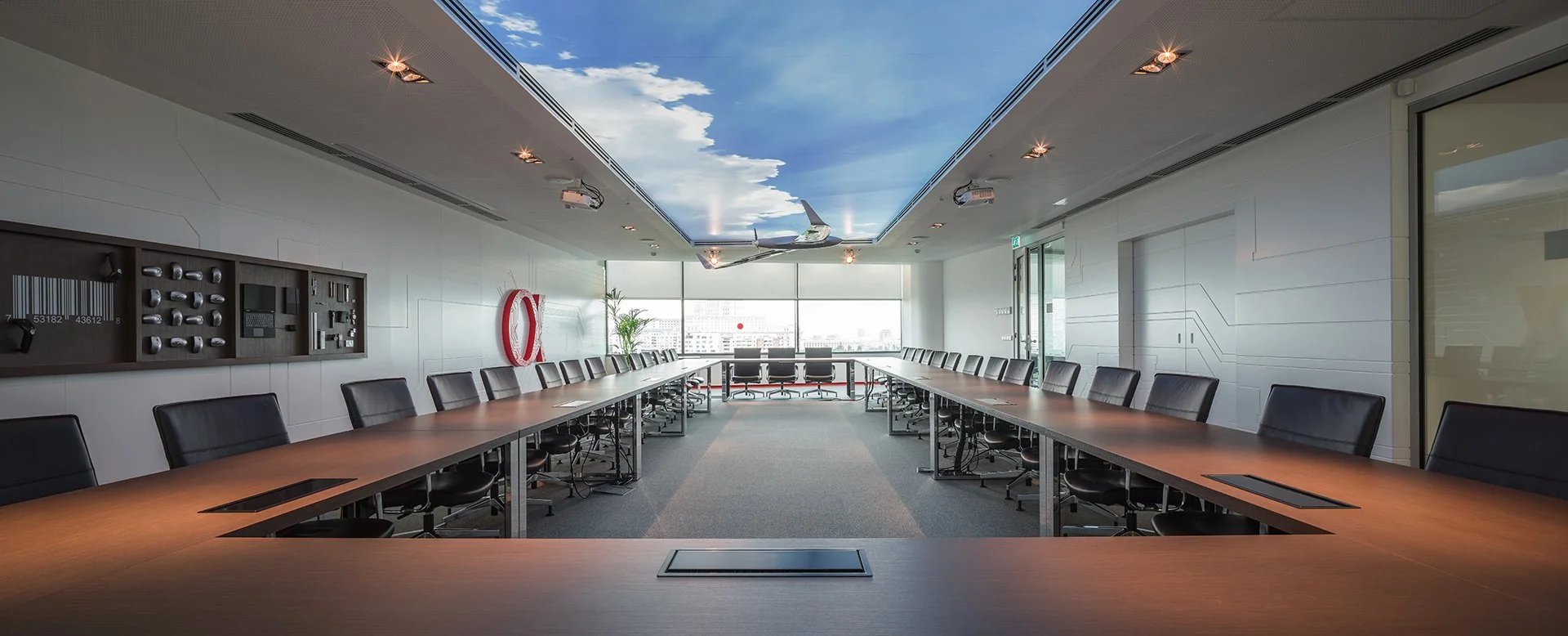 A modern conference room with a U-shaped wooden table, black leather chairs, a blue sky with clouds mural on the ceiling, and a city view through large windows.