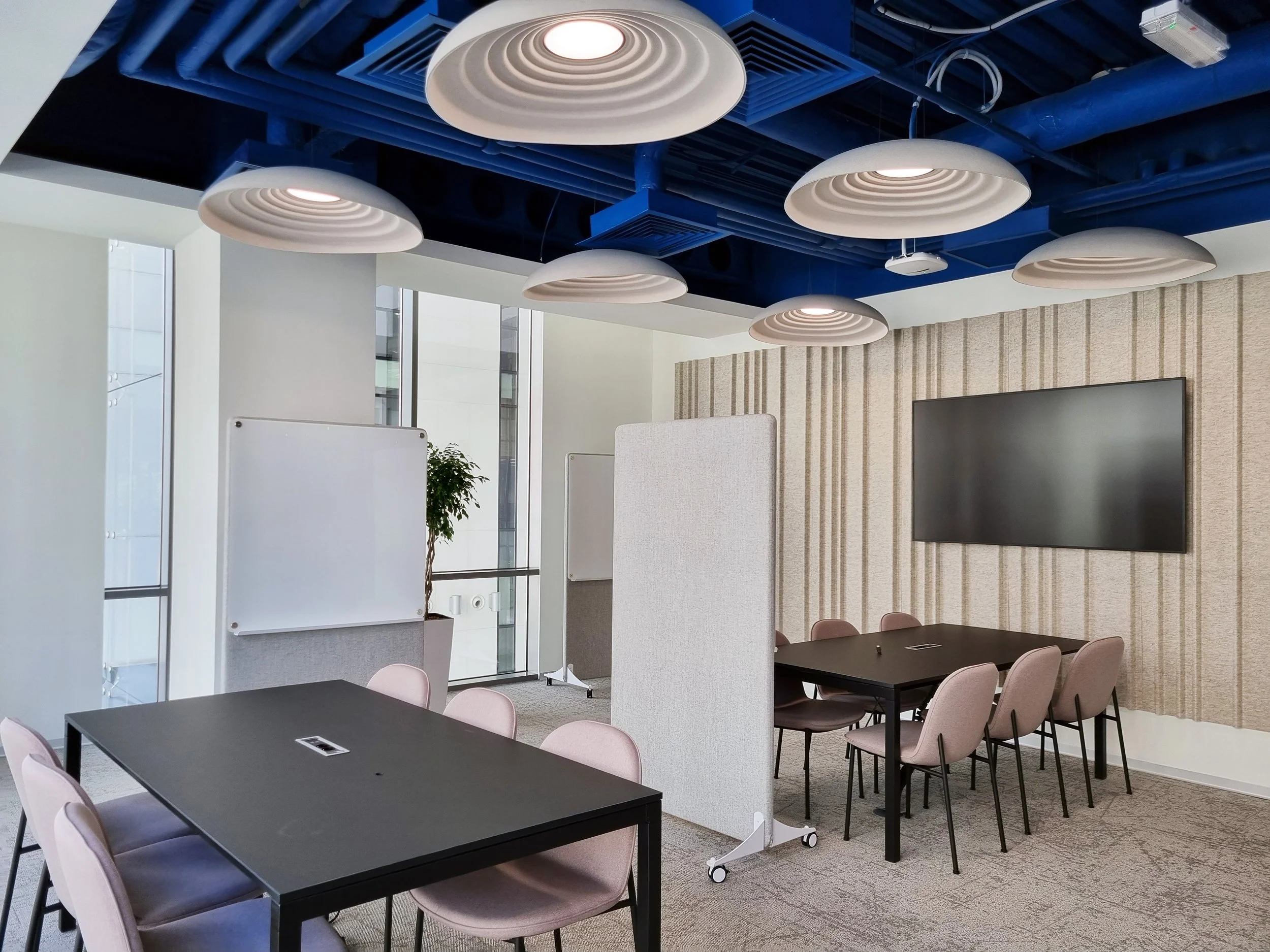 Modern conference room with black tables, pink chairs, whiteboards, a large wall-mounted TV, and a split privacy screen. The ceiling features blue and beige circular light fixtures.