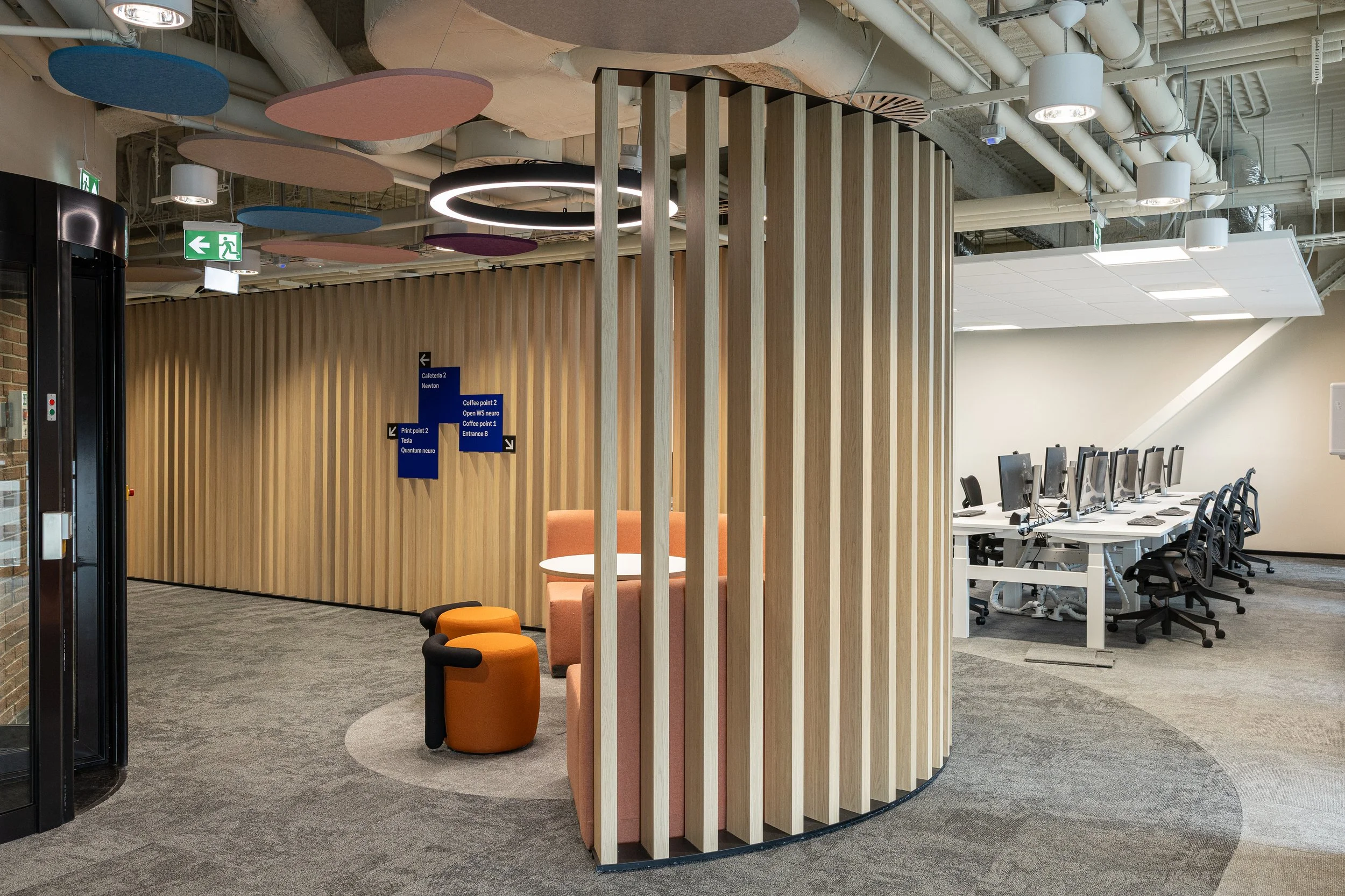 Modern office interior with a curved wooden slat partition, seating area with pink and orange chairs, and a row of desks with computer monitors in the background.