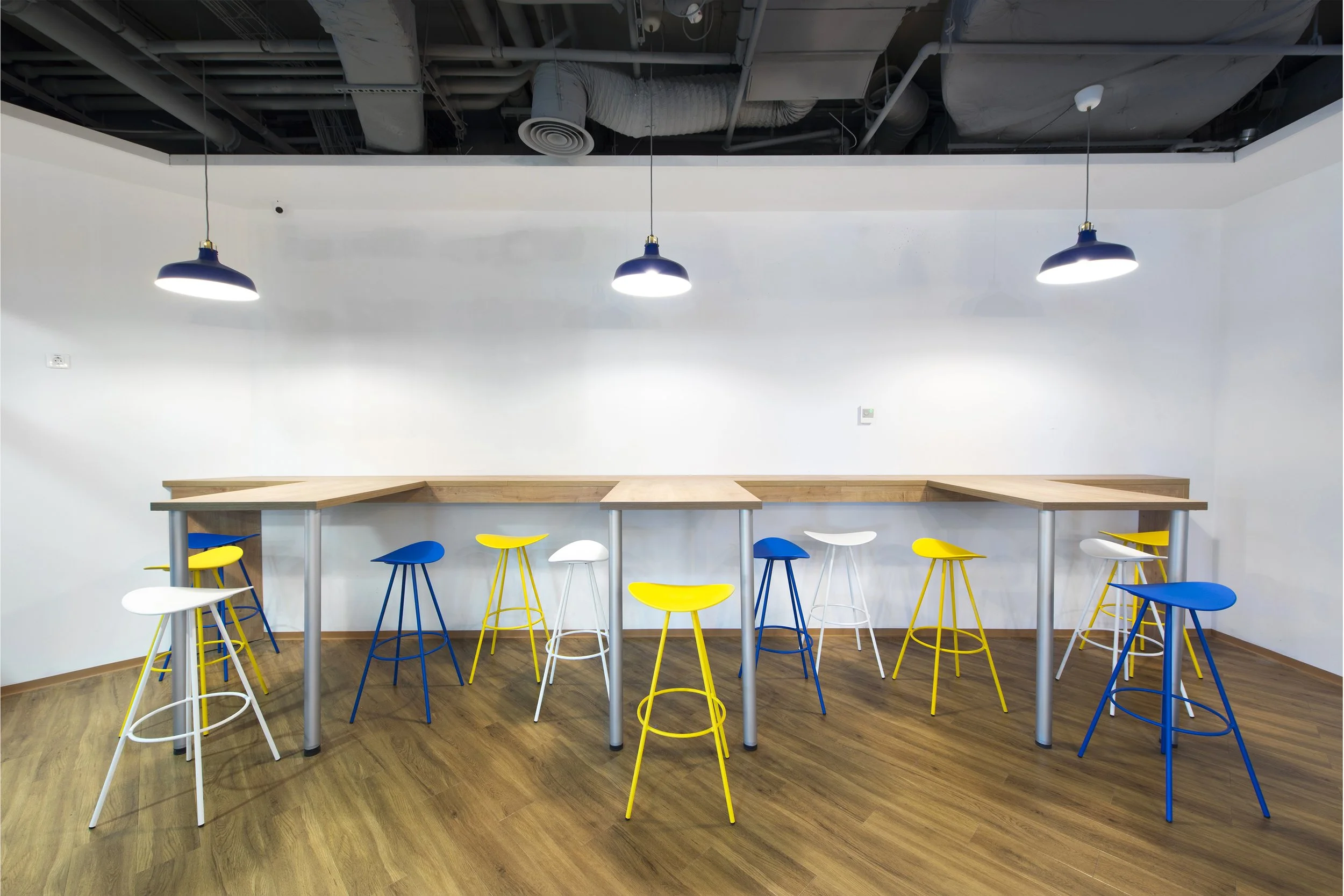 Empty room with wooden flooring, white walls, three hanging black lamps, a long high wooden table, and colorful barstools in blue, white, and yellow.