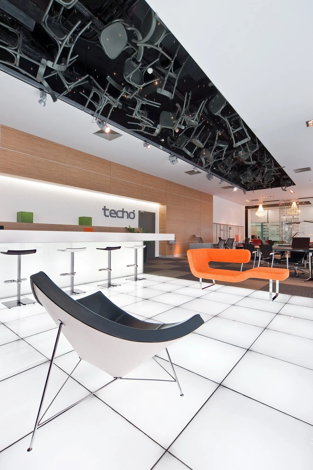 Modern office lobby with illuminated white tiled floor, contemporary black and orange chairs, and a reception desk with 'techo' sign. Ceiling features a unique installation of stacked chairs and black panels.