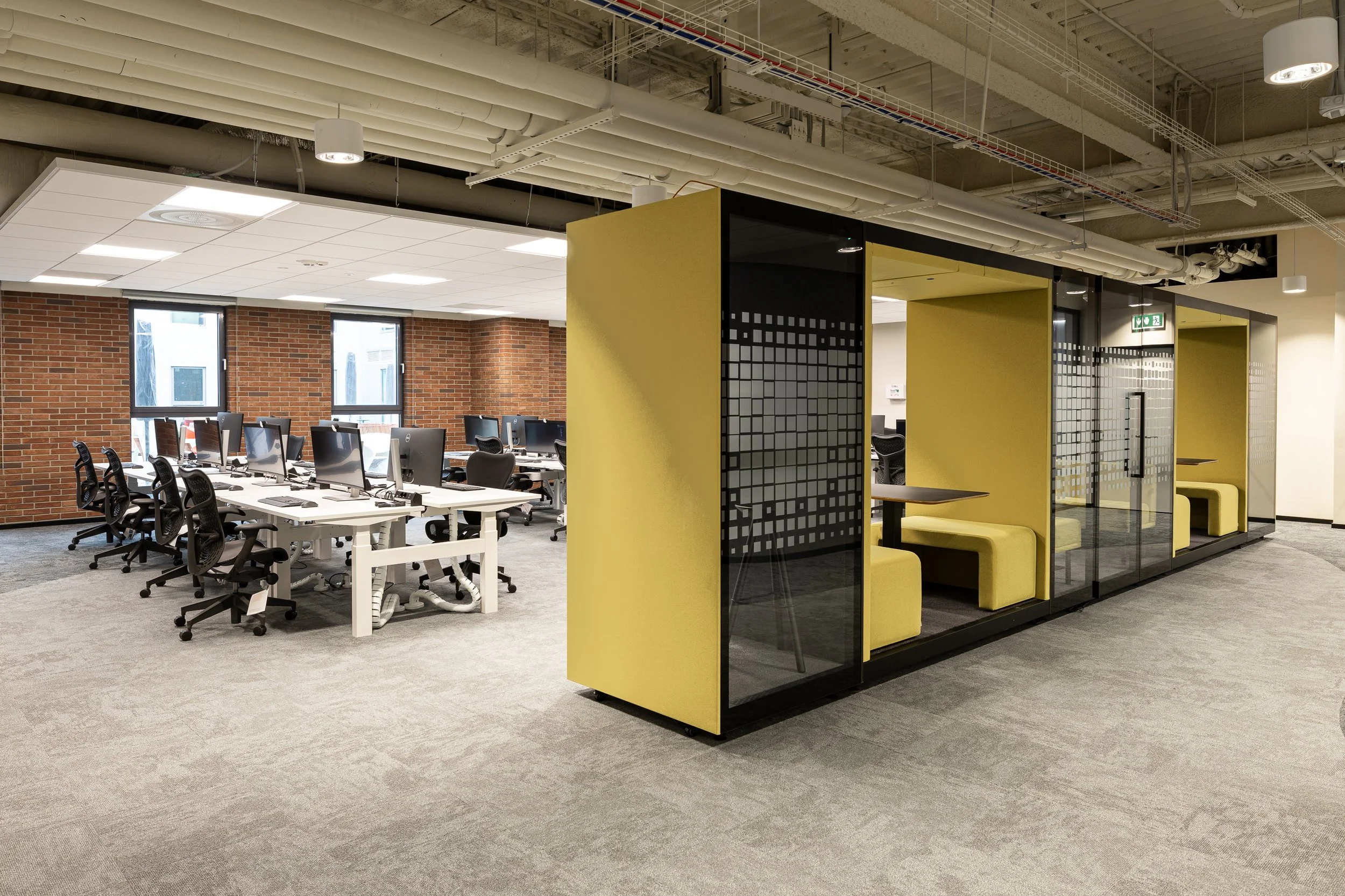Open office workspace with desks, computer monitors, and chairs, including a semi-private seating area with yellow booths and glass walls.