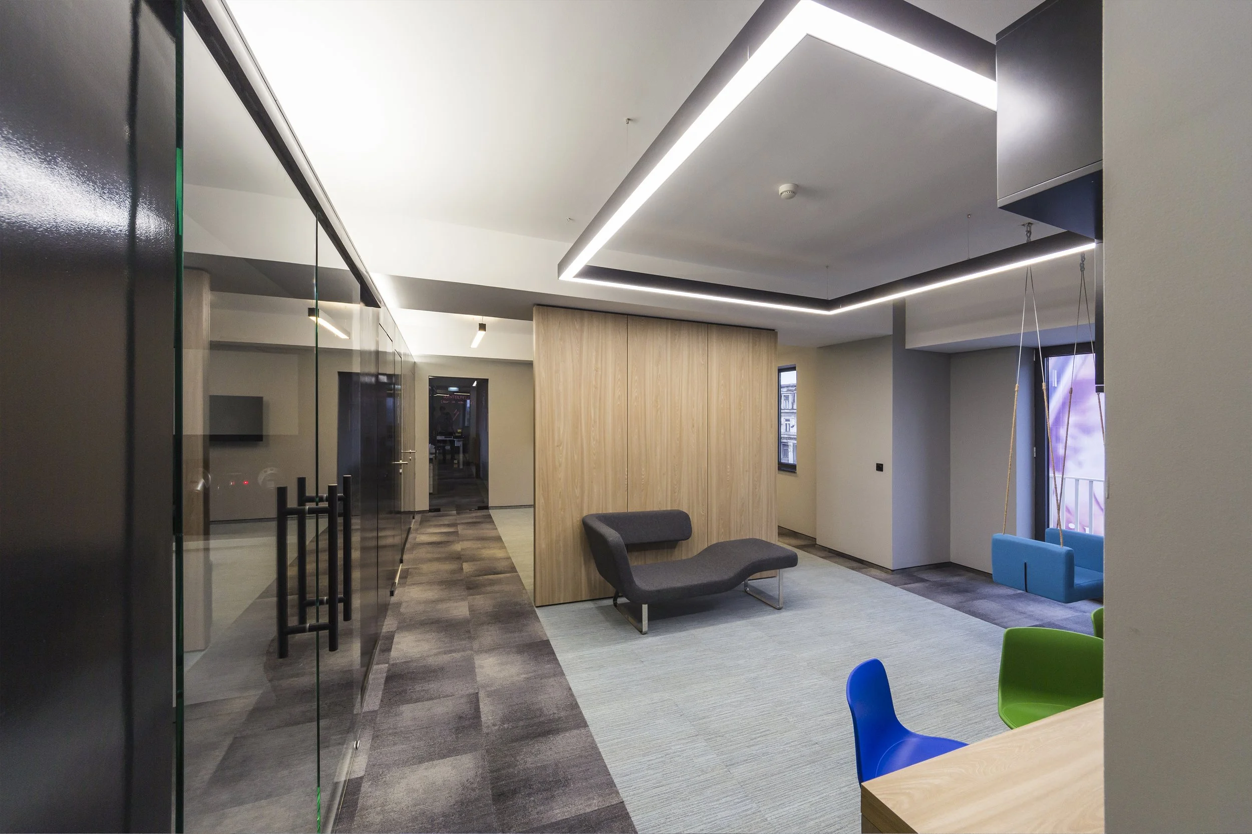 Modern office lounge area with a black sofa, colorful chairs, wooden wall, and suspended blue seating. Overhead lighting and windows allow natural light.