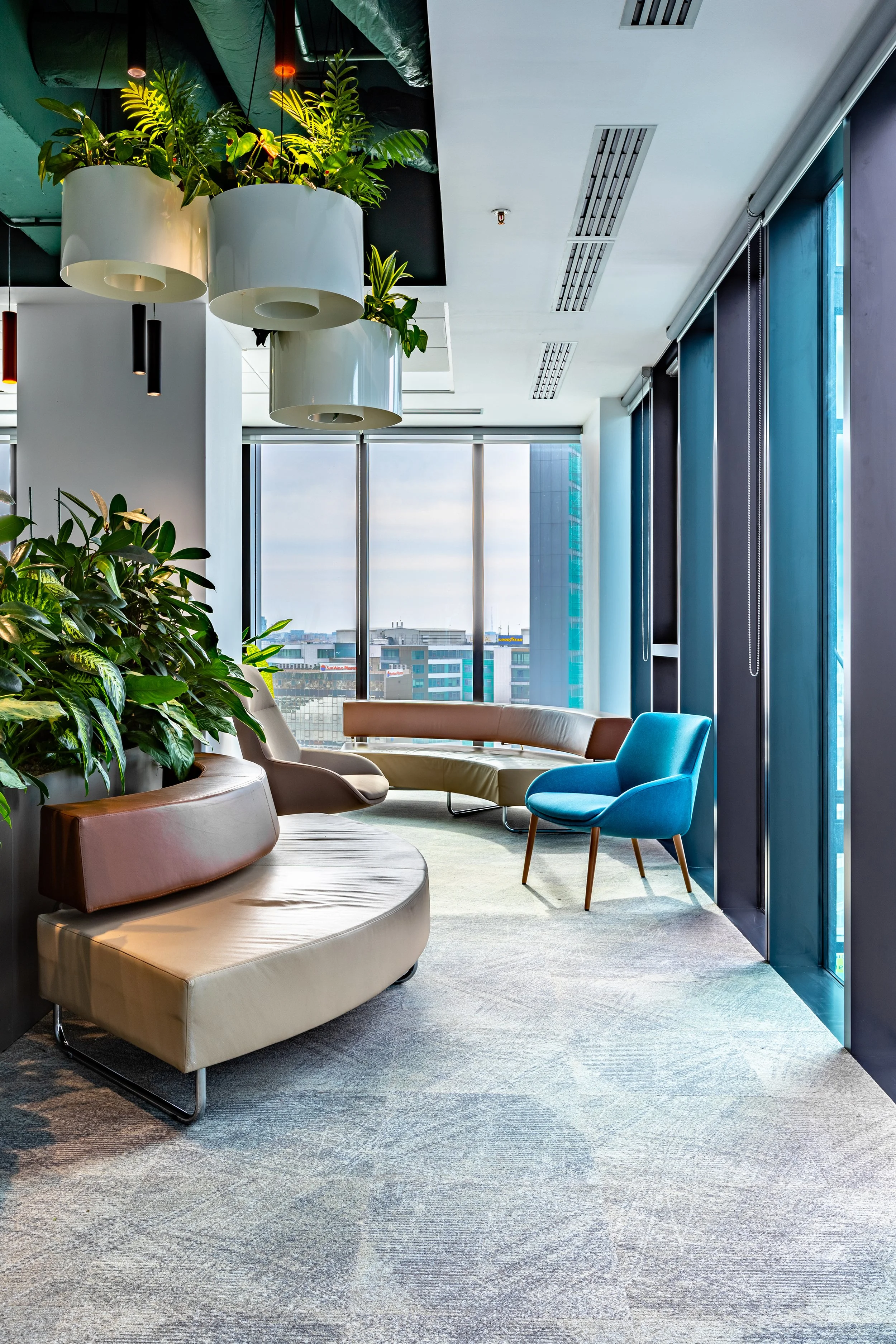 Modern office lounge area with colorful chairs, green plants, large windows, and cityscape view.