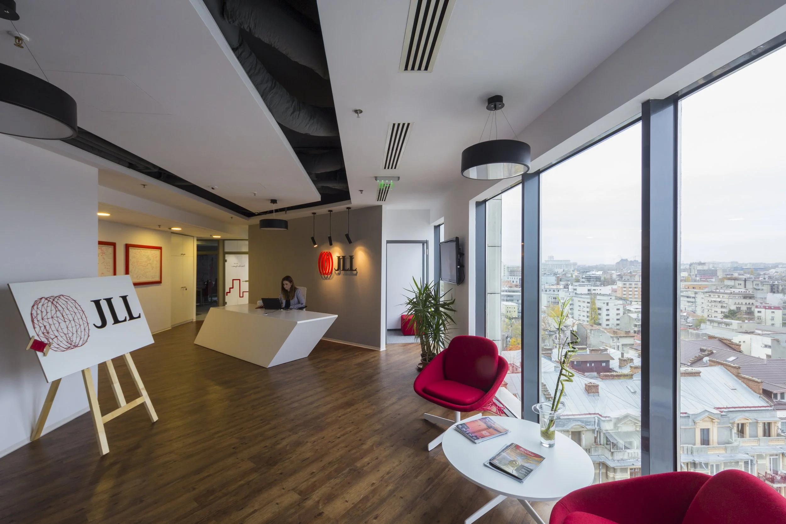 Interior of an office with a reception desk, red chairs, potted plants, large windows showing a cityscape, and branded JLL signage.