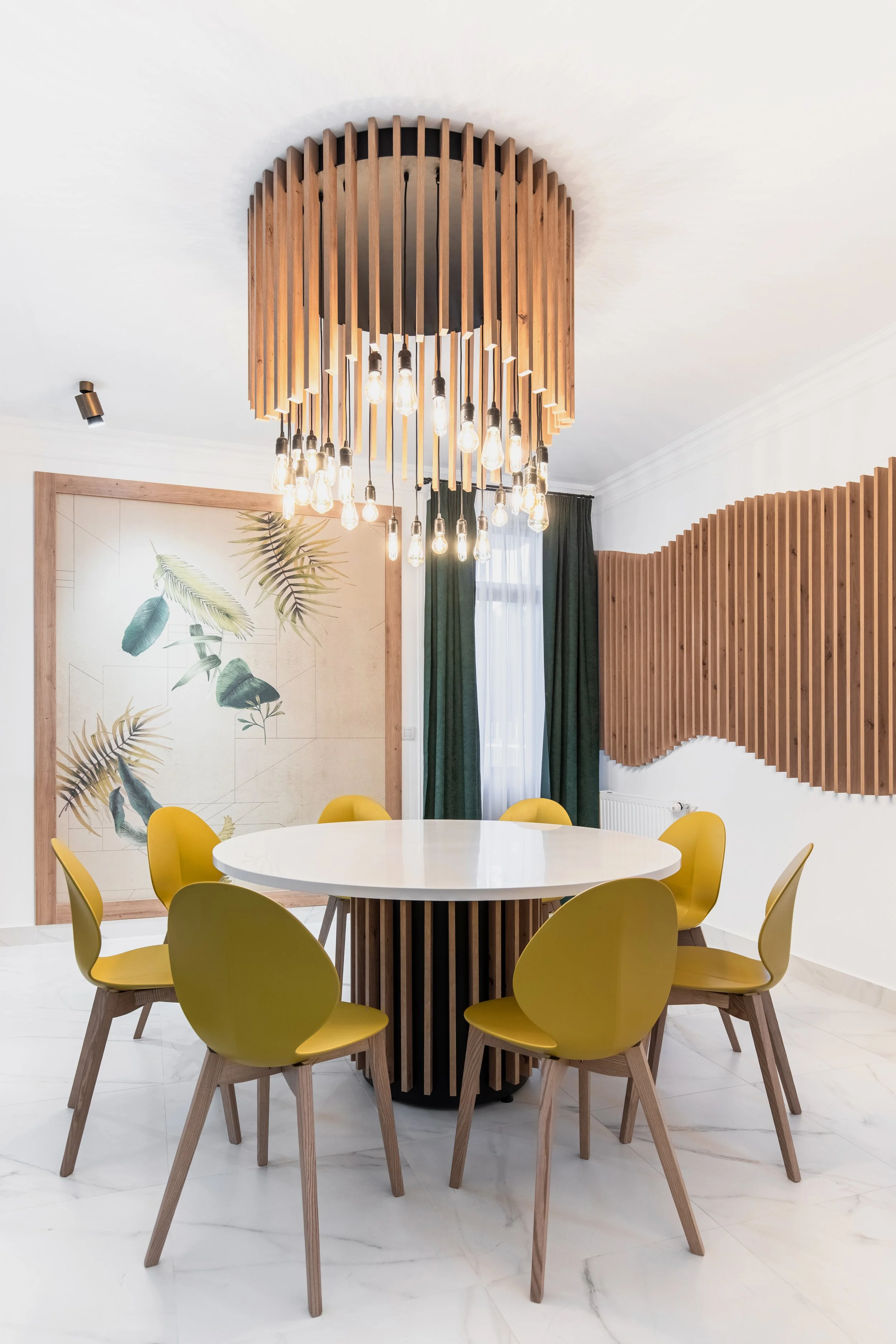 Modern dining room with a white round table, six yellow chairs with wooden legs, a wooden decorative wall, and a large wooden chandelier with exposed light bulbs.