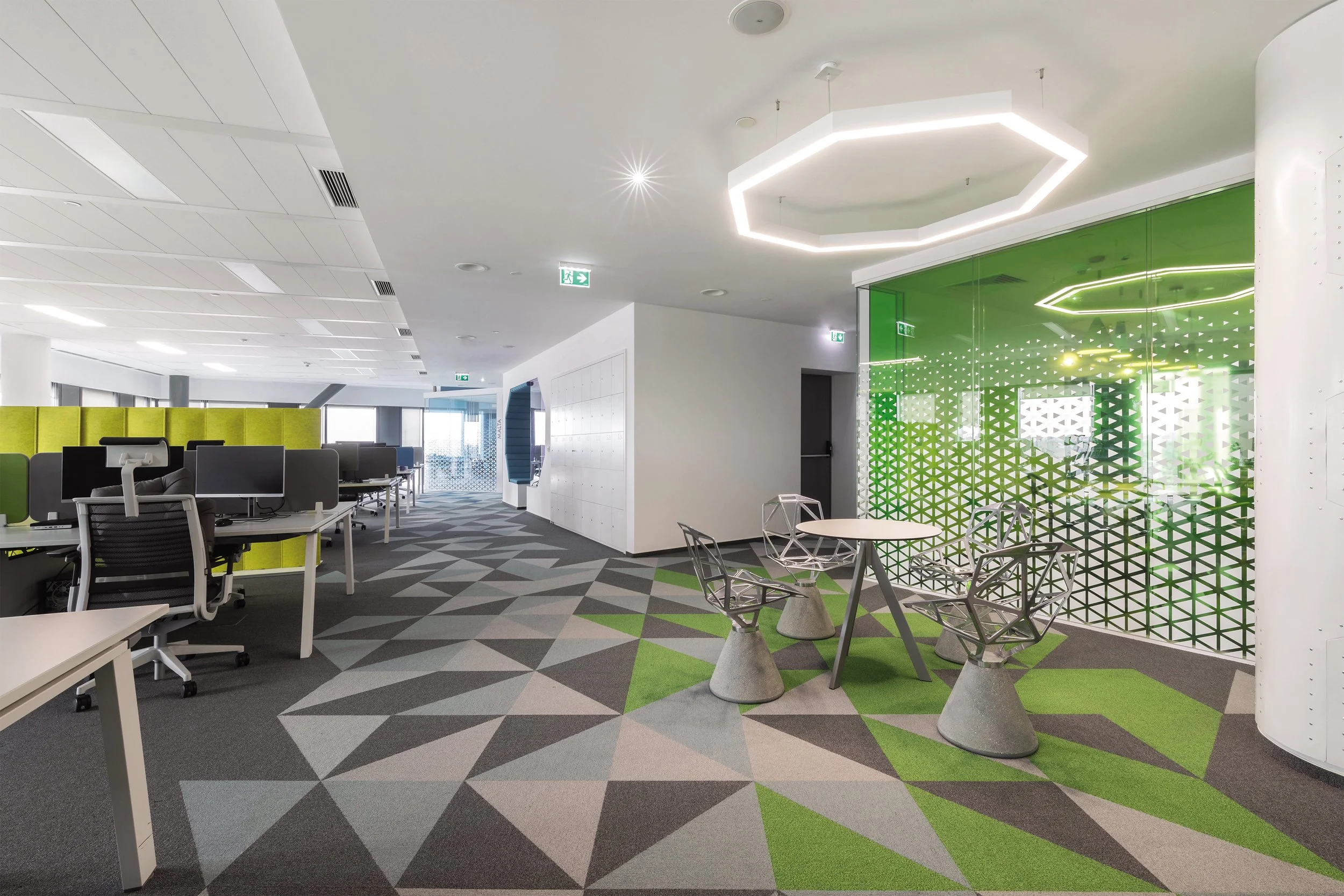 Modern office space with workstations, a green accent wall with geometric pattern, a small round table with four modern chairs, and geometric patterned carpet.