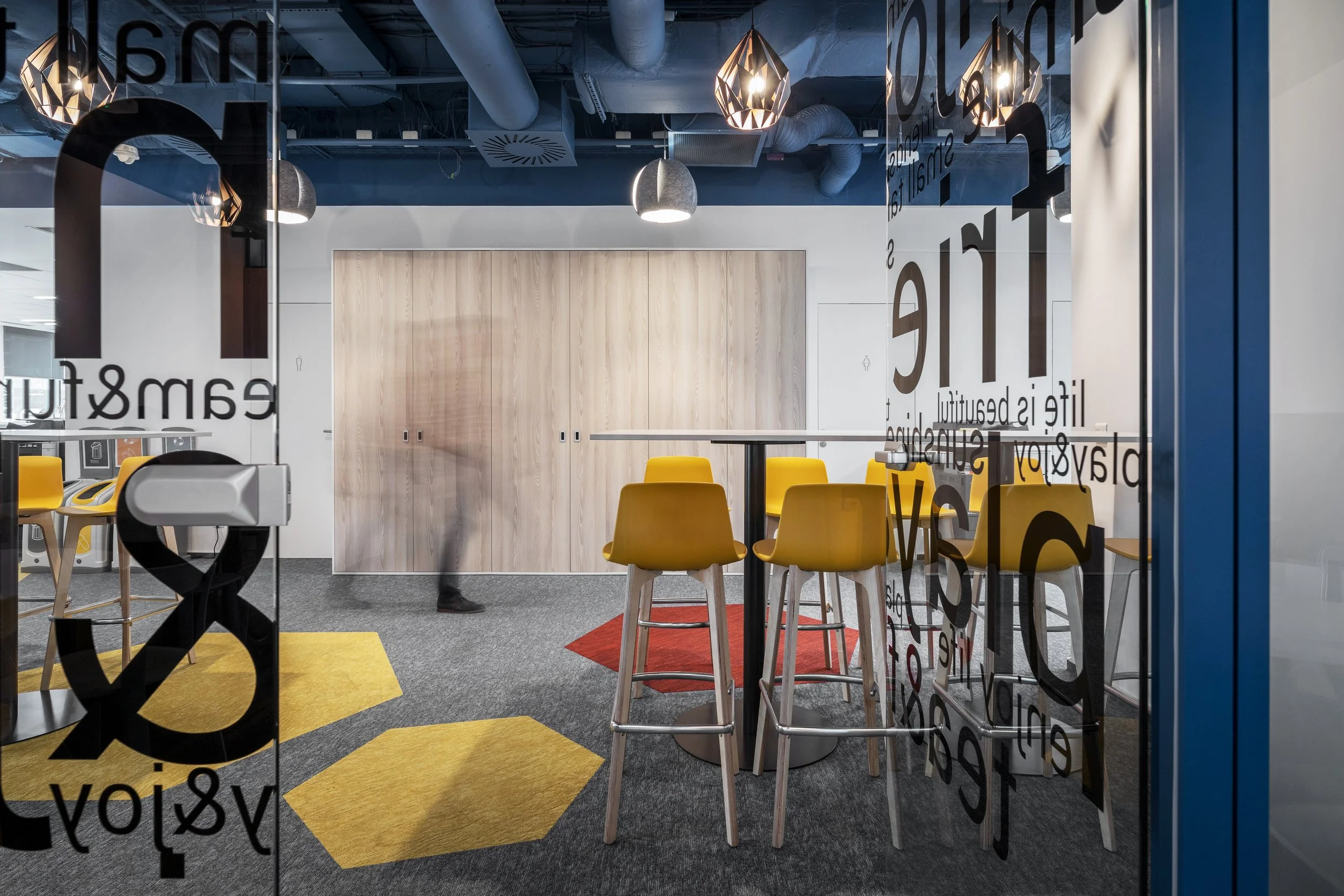 Modern office lounge area with yellow bar stools around high tables, gray carpet flooring with yellow hexagon rugs, wooden lockers, and decorative hanging lights. The photo is taken through a glass wall with black text.