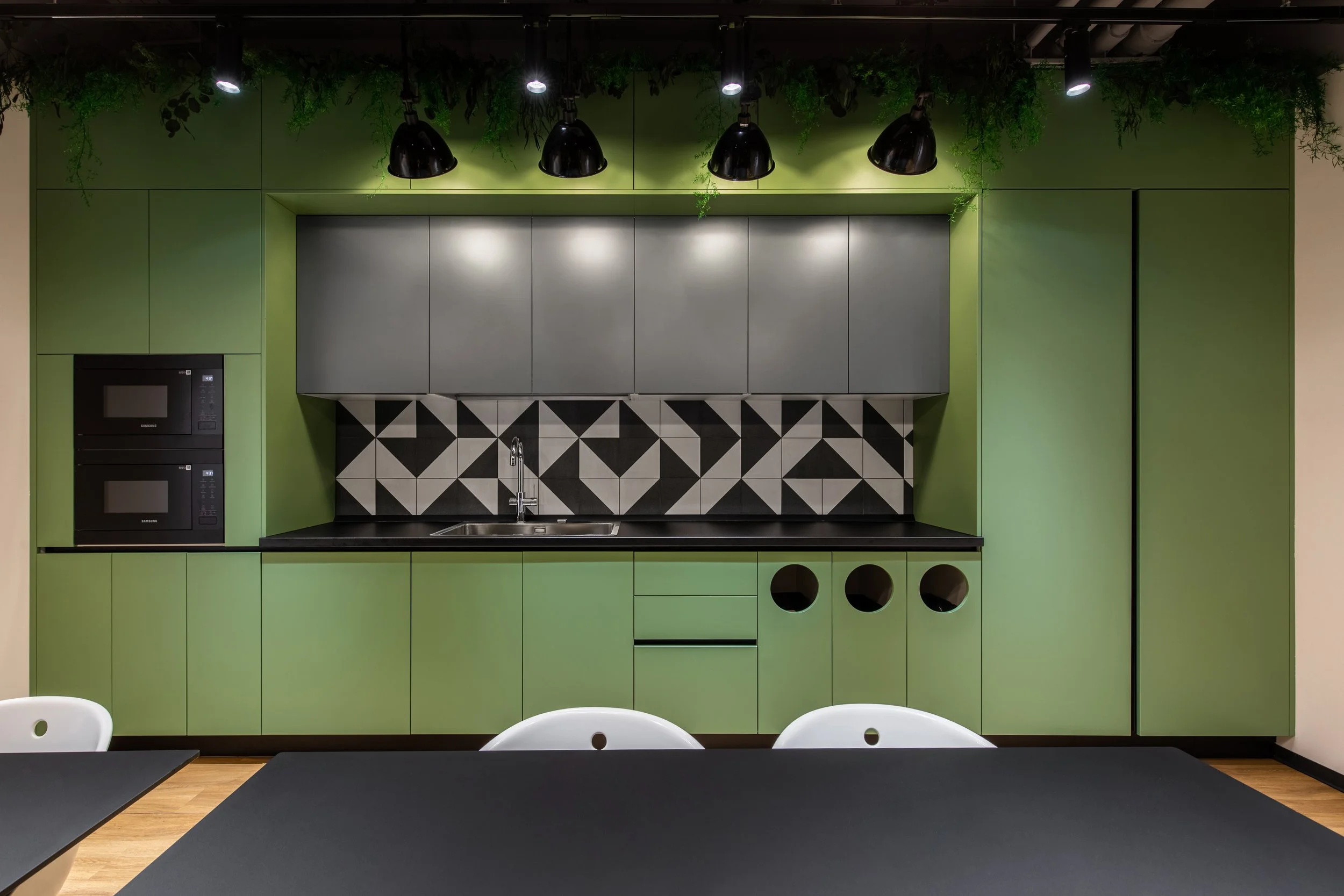 Modern kitchen with green cabinets, black countertop, geometric patterned backsplash, stainless steel sink, and black spot cabinet openings, with black ceiling lights and green ceiling decor.