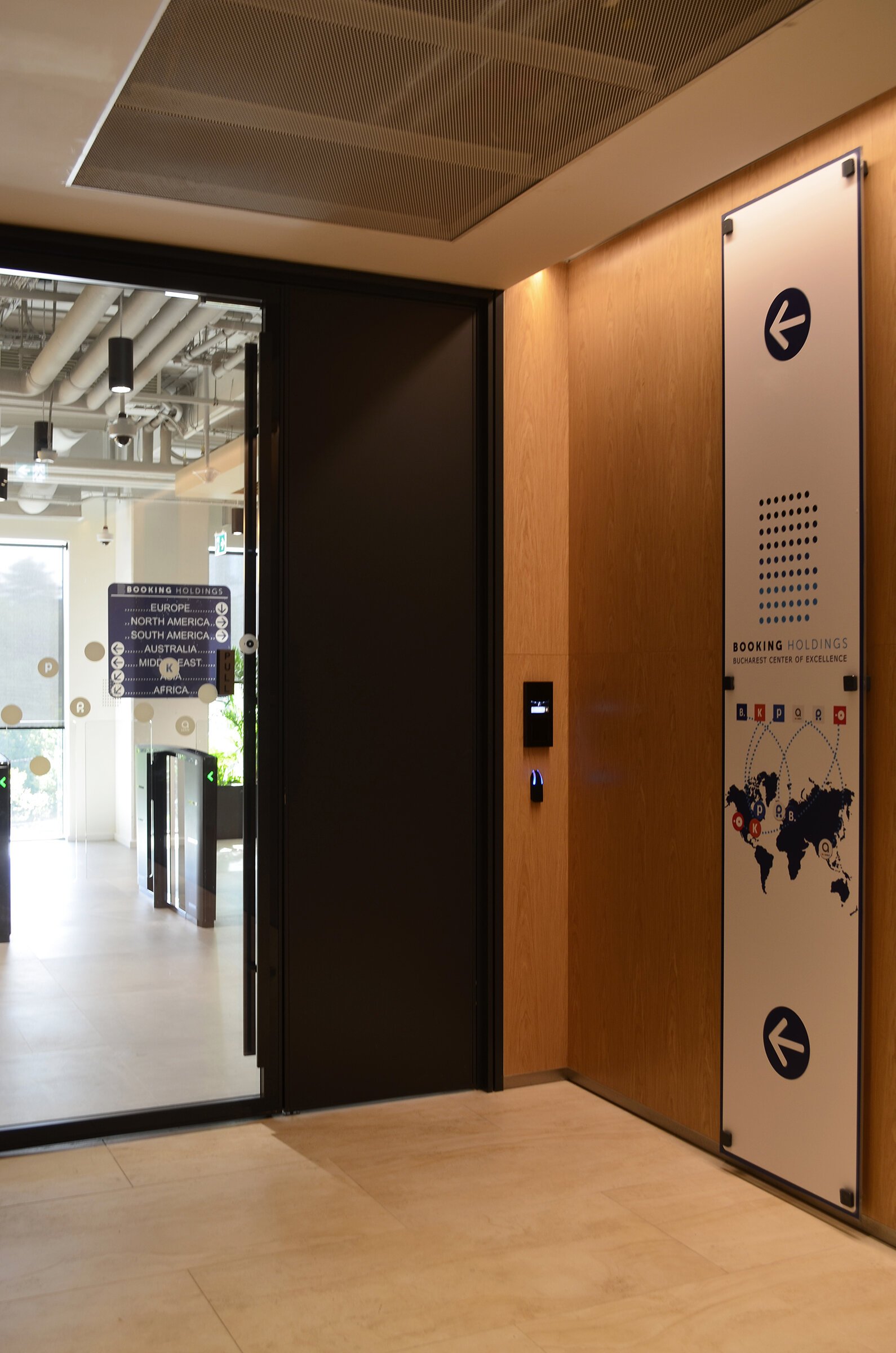 Interior view of a building with wooden wall panels, an automated door, and a large sign with a world map and walking directions to different regions. The sign indicates booking holdings.