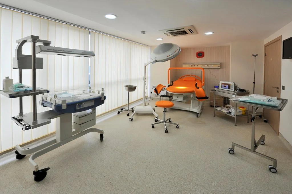 Room with medical equipment, including a hospital bed, incubator, radiology machine, and a hospital bed with orange accents, in a medical setting.