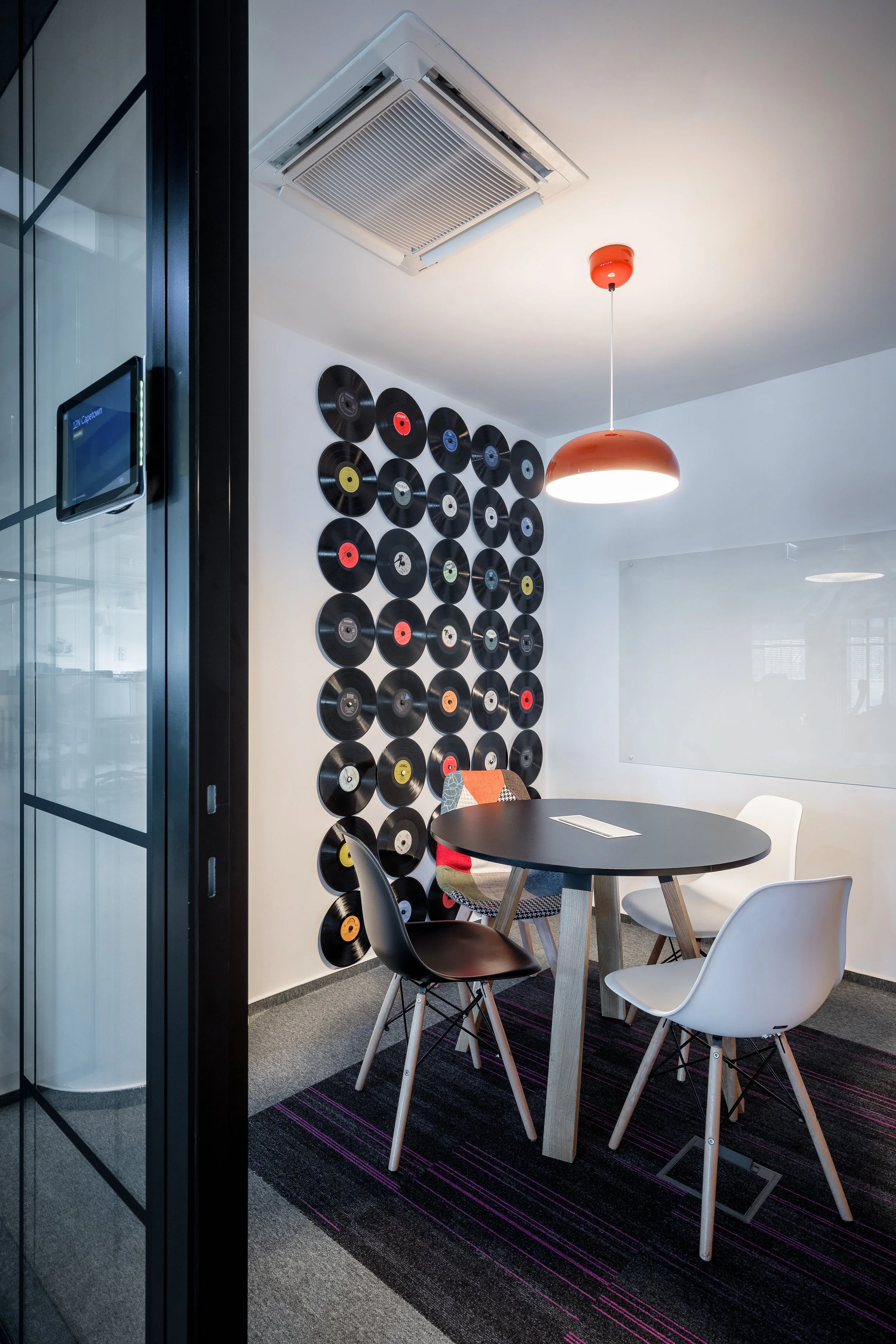 A modern meeting room with a round black table surrounded by three chairs with different designs. The wall behind features a decorative art installation of vinyl records. There is a white ceiling with a ceiling-mounted air conditioning unit and a hanging orange pendant light. The floor has a dark rug with purple accents.
