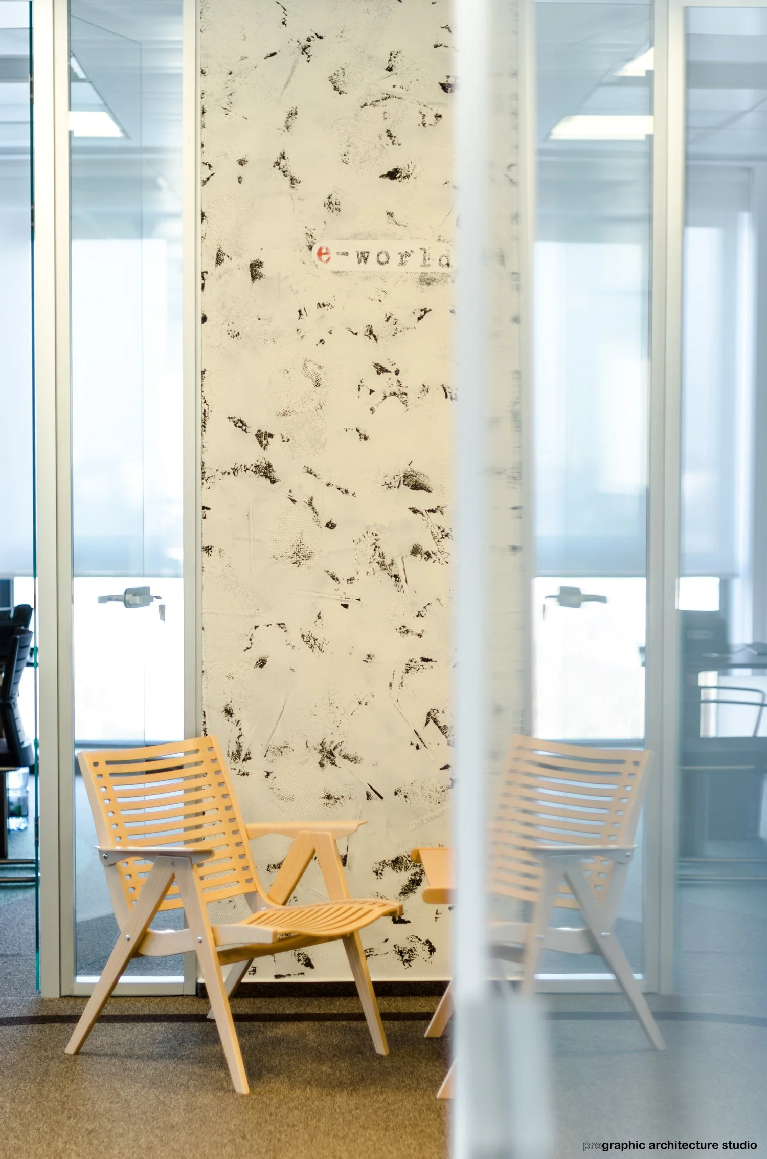 Two wooden chairs with slatted backs and seats, positioned in front of a textured white wall with black markings, inside a modern office space with glass partitions and windows.