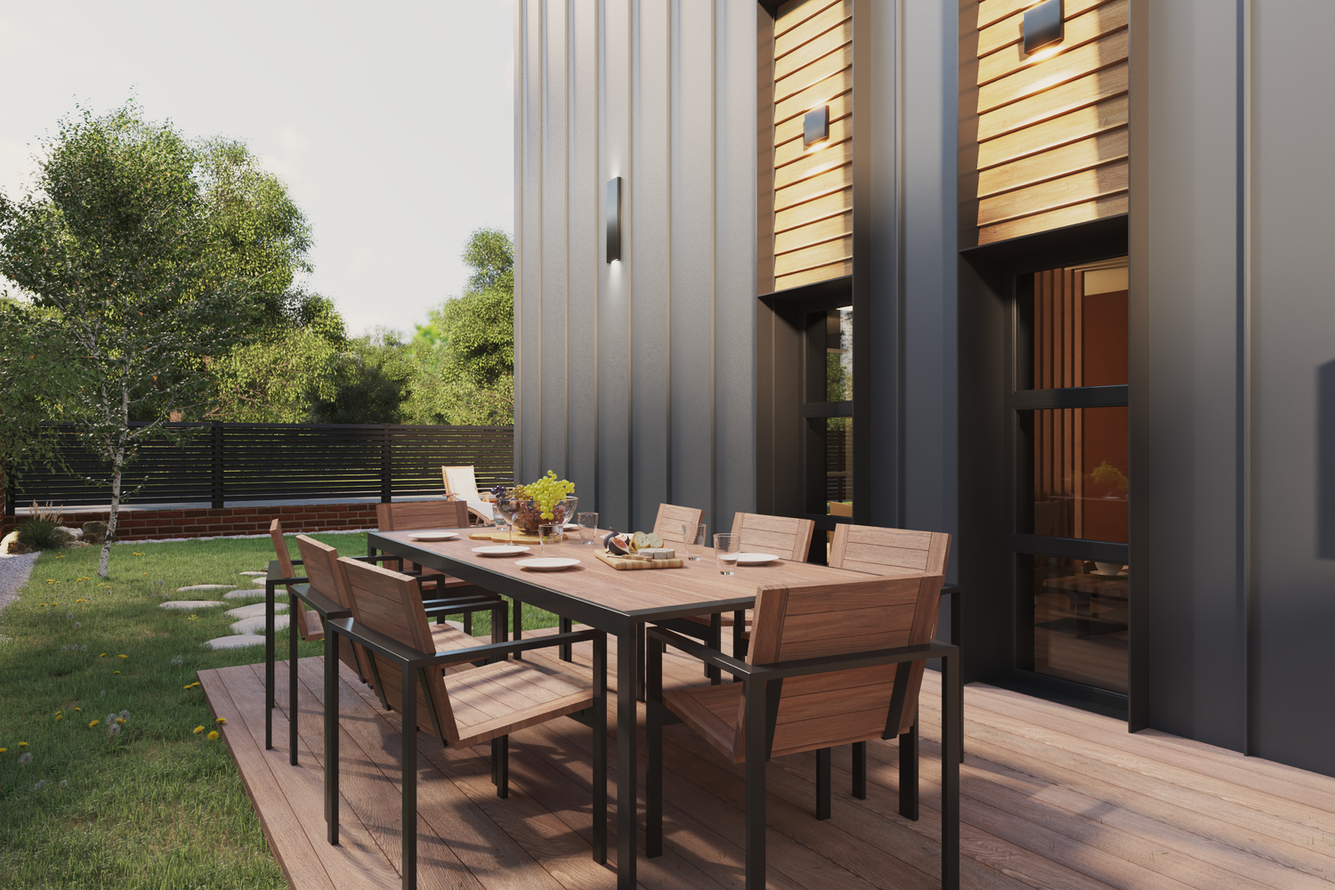 Outdoor dining area with a wooden table and chairs set on a deck, overlooking a grassy backyard with trees, a black fence, and modern exterior wall with wood and metal siding.