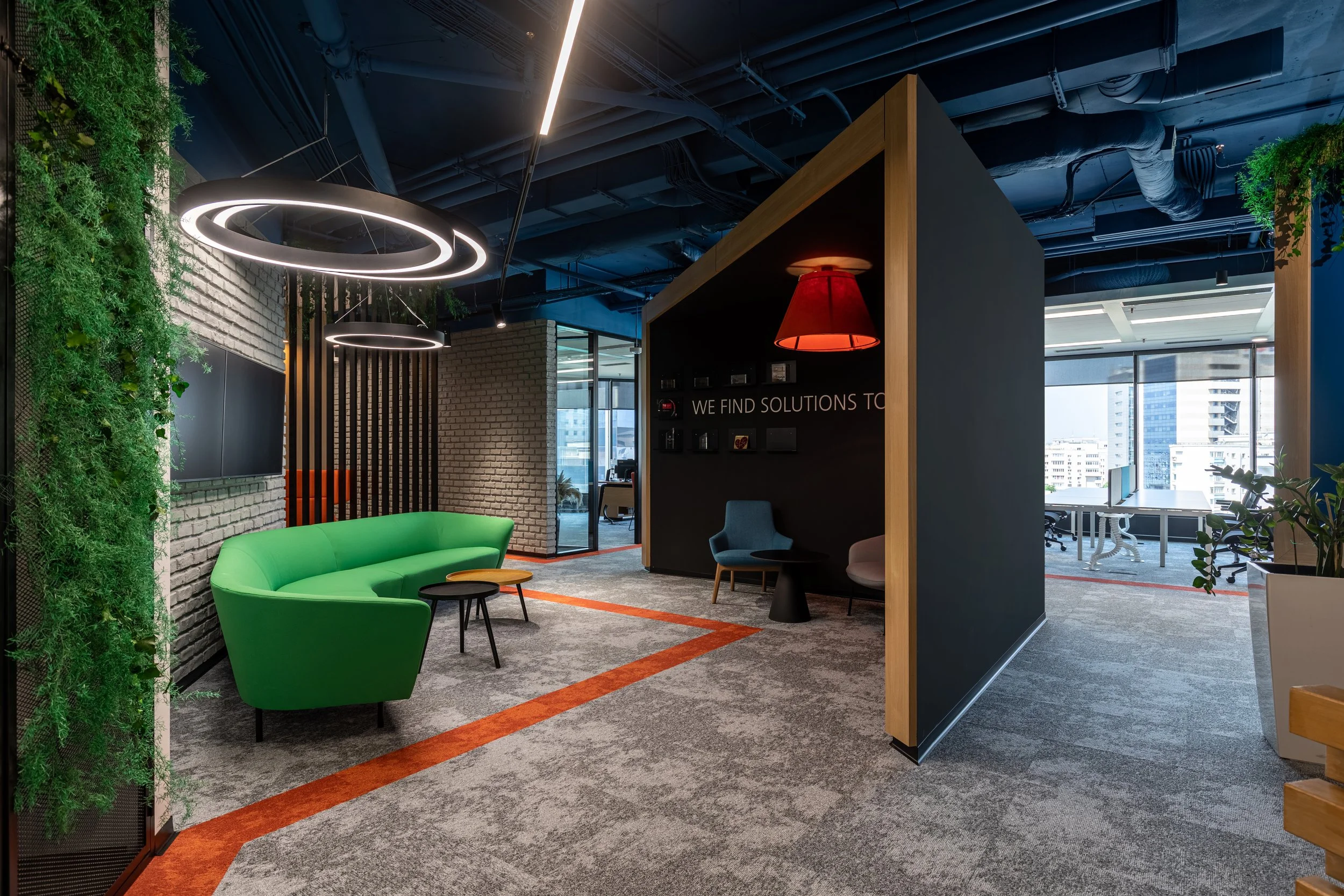Modern office lounge area with green sofa, black tables, hanging circular light fixtures, textured white brick walls, a black wall with the partially visible phrase 'WE FIND SOLUTIONS TO', and large windows showing cityscape.