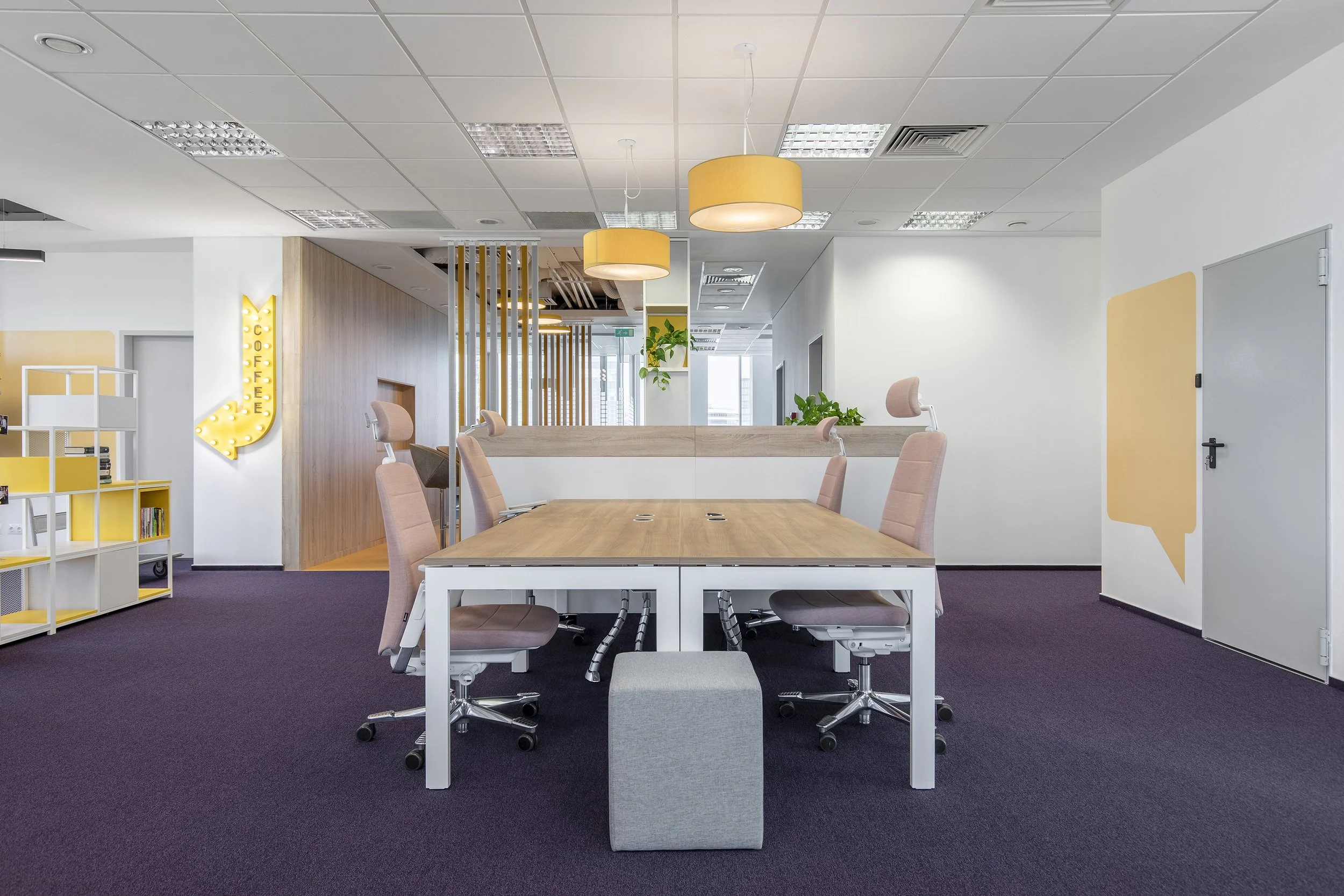 Modern office meeting room with a wooden table surrounded by eight pink office chairs. The room features hanging yellow pendant lights, a purple carpet, white walls with yellow speech bubble and arrow decor, plants, and a partial view of a bookshelf with yellow accents.