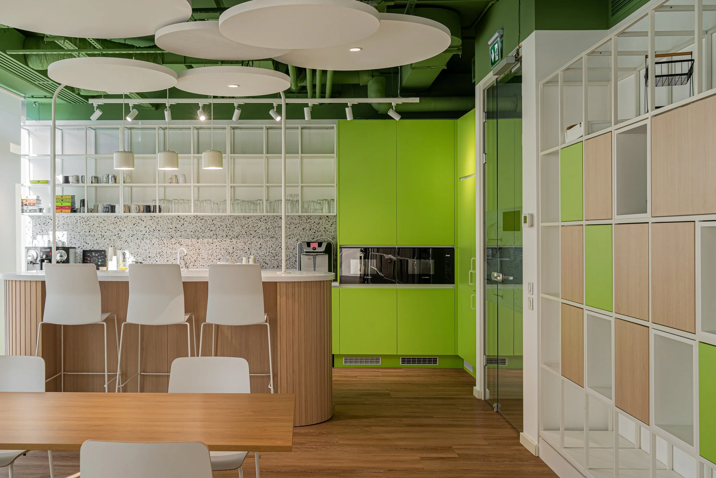 Modern kitchenette area with white chairs, wooden table, green cabinets, and open shelving with cups and glasses, in an office or cafe setting.