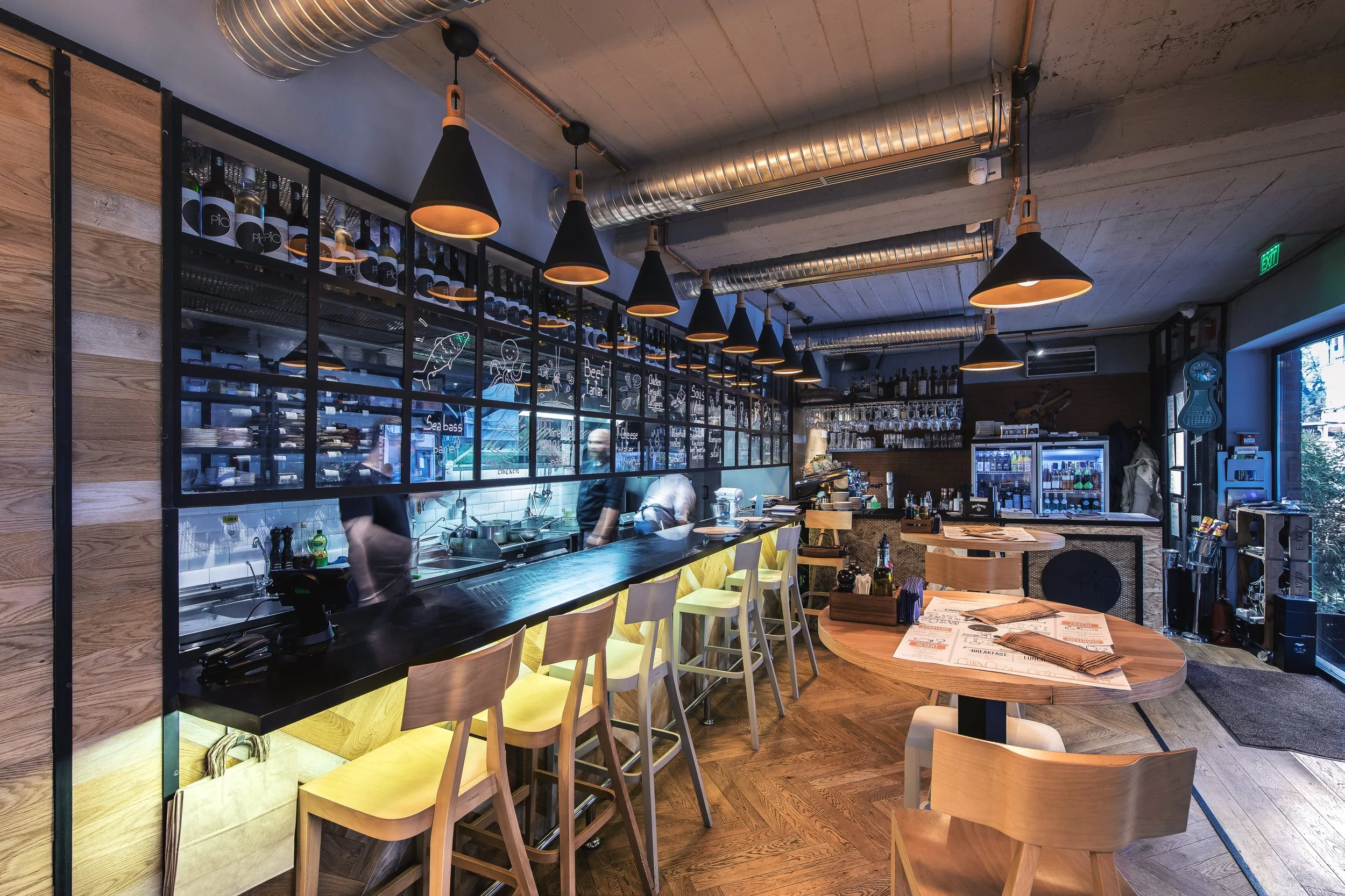 Interior of a modern restaurant with a bar counter, high chairs, and tables, decorated with wood and black accents, hanging pendant lights, and a large window letting in natural light.