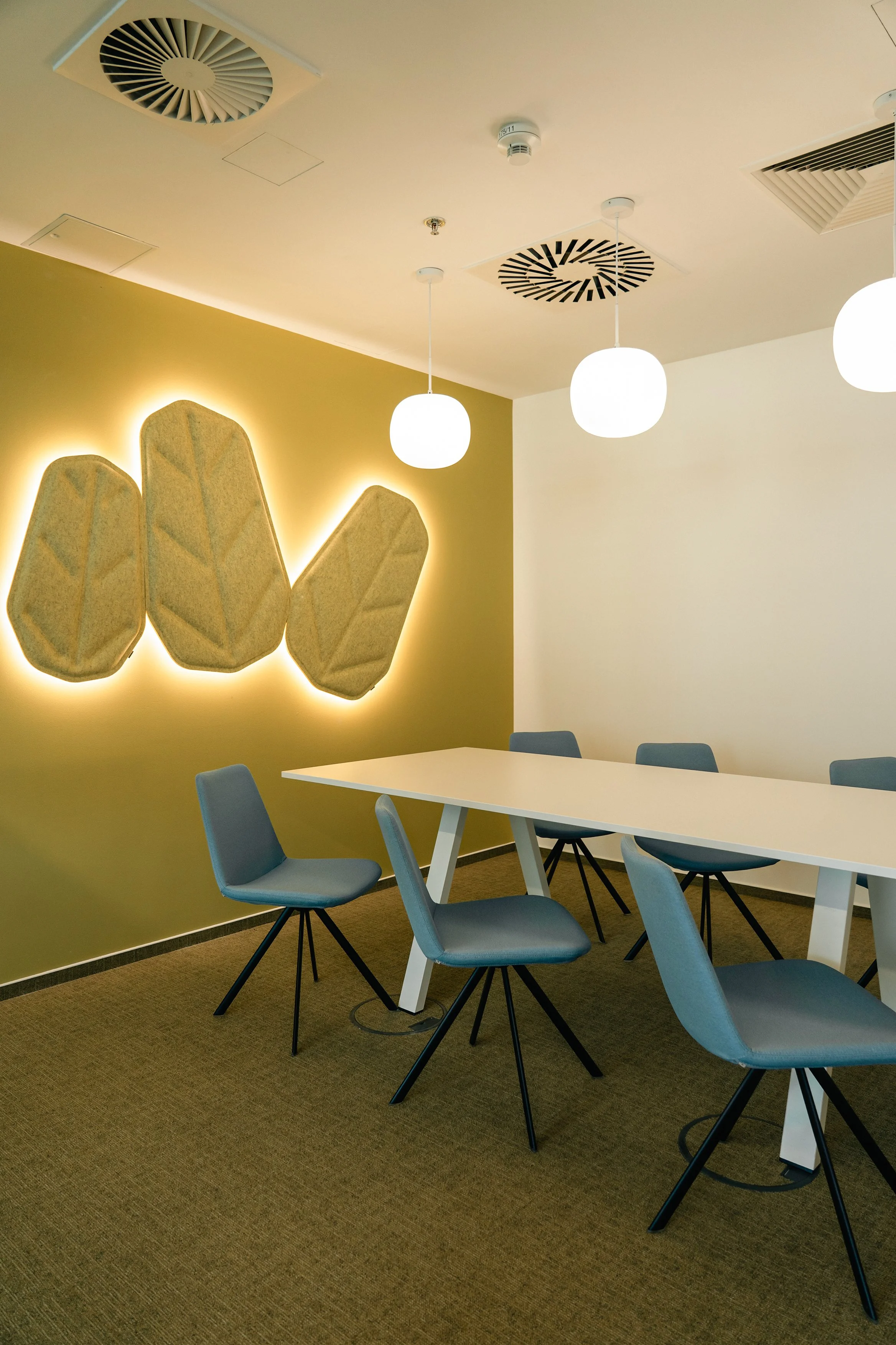 A modern conference room with a long white table surrounded by gray chairs on black splayed legs. The wall on the left side has a large decorative art piece resembling leaves, with soft backlighting. The ceiling features circular light fixtures and air vents.