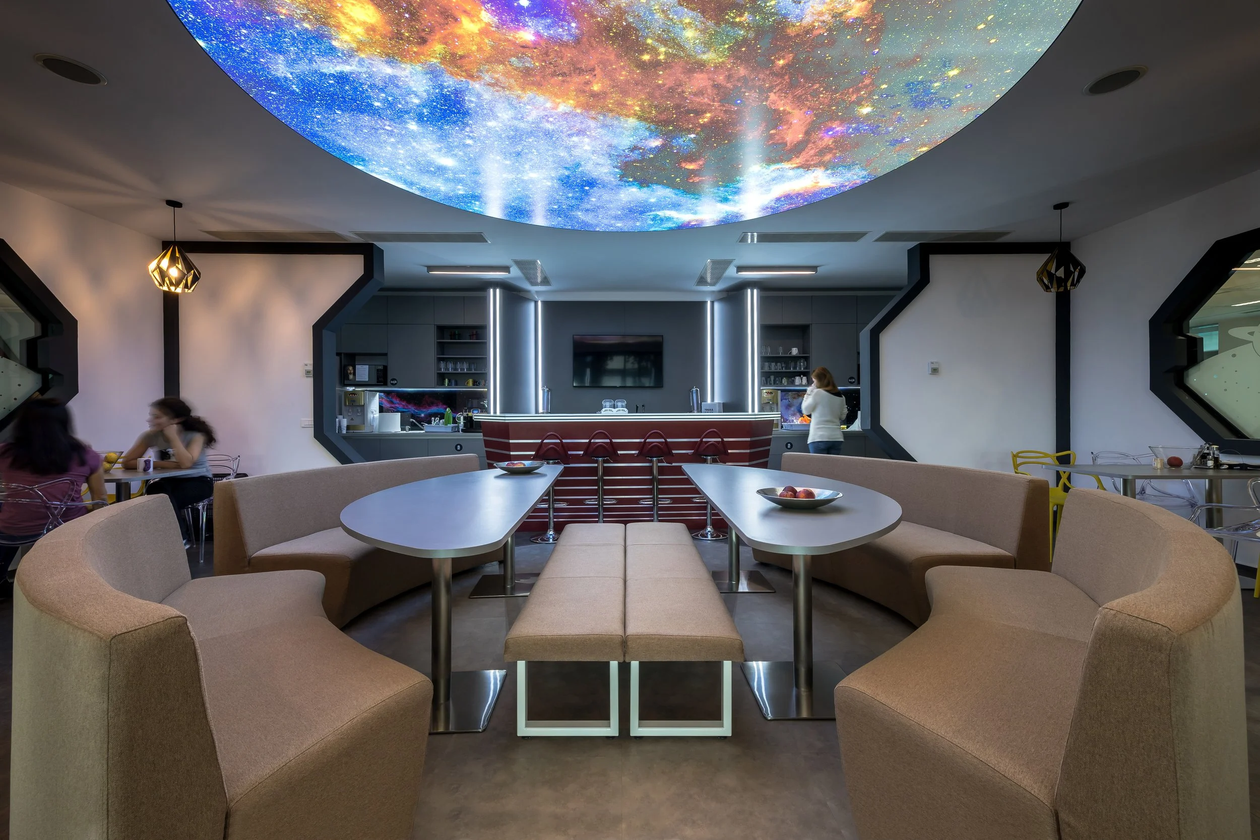 Modern lounge or restaurant with curved beige booth seating around white tables, a bar with a red-striped front, and a ceiling with a large, colorful starry sky projection. Two women are sitting at a table on the left, and another woman is at the bar on the right. There are some fruit bowls on the tables and ambient lighting.