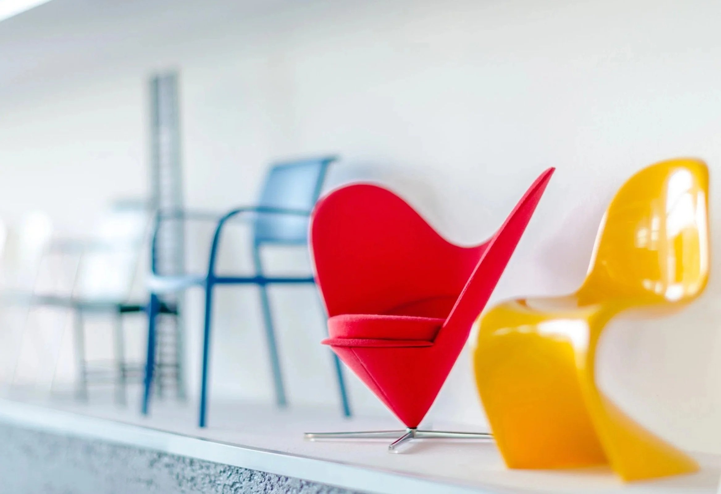 Colorful modern chairs arranged in a row on a shelf, with a focus on a red chair shaped like a checkmark and a yellow chair with a curvy design, against a white wall.