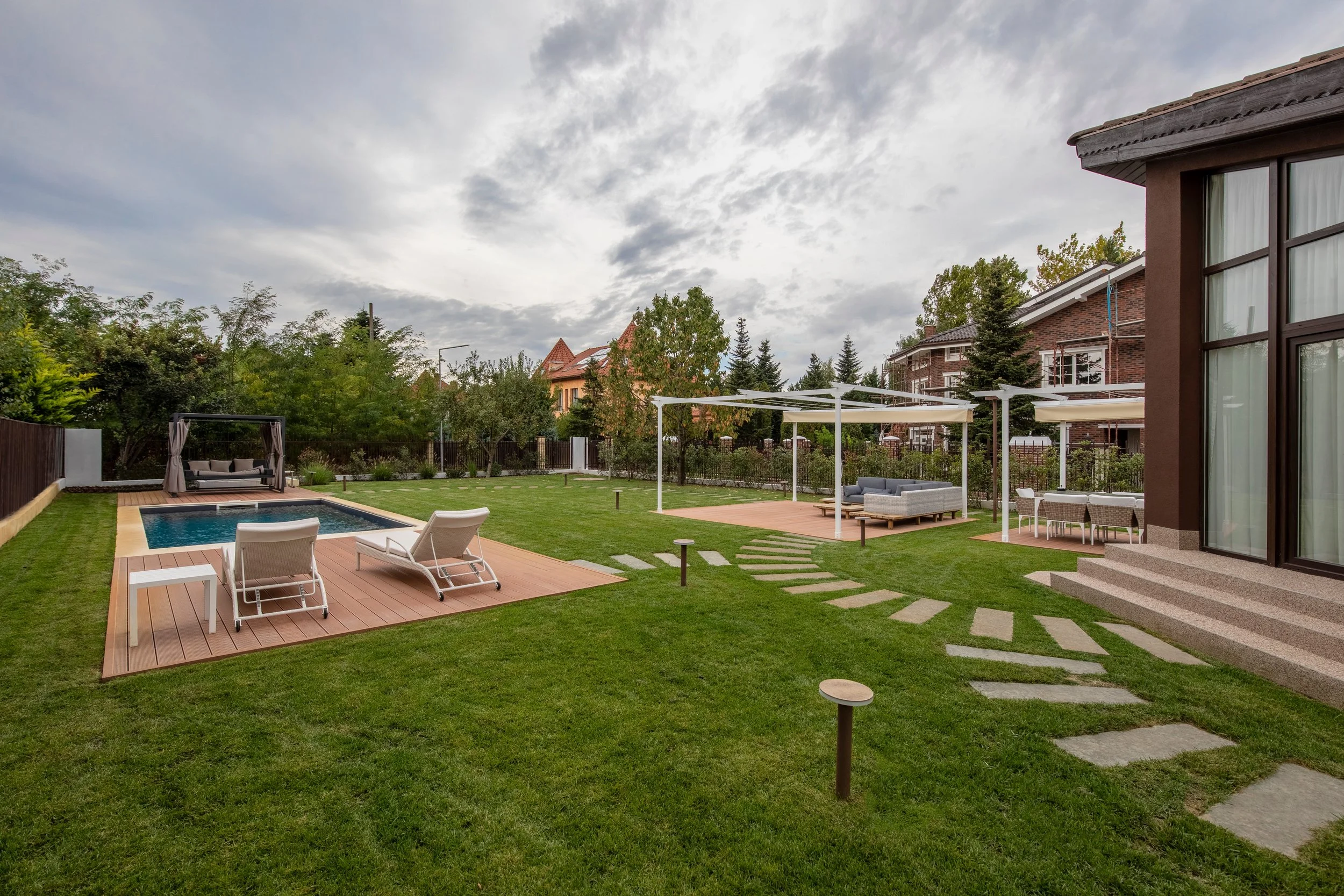Spacious backyard with a swimming pool, sun loungers, a shaded seating area, a stone pathway, and a well-maintained lawn under cloudy skies.