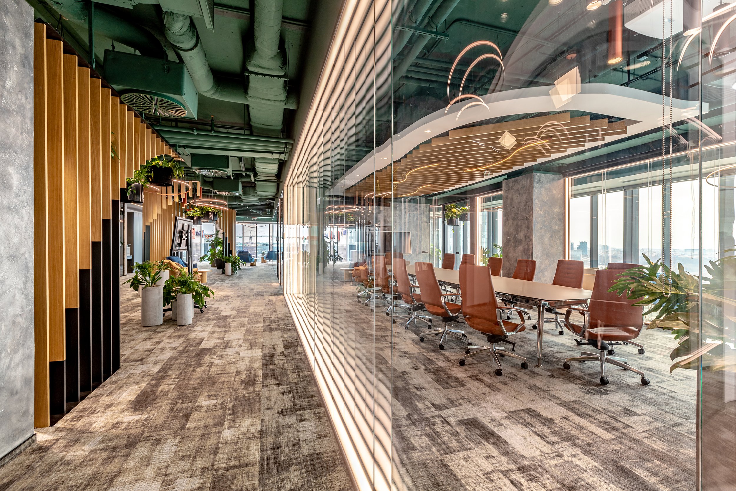 Modern office interior with a glass-walled conference room on the right side, featuring brown chairs and a long white table. The hallway on the left side has wooden slats, potted plants, and a carpeted floor. The ceiling has exposed green pipes and ducts, with contemporary lighting fixtures.
