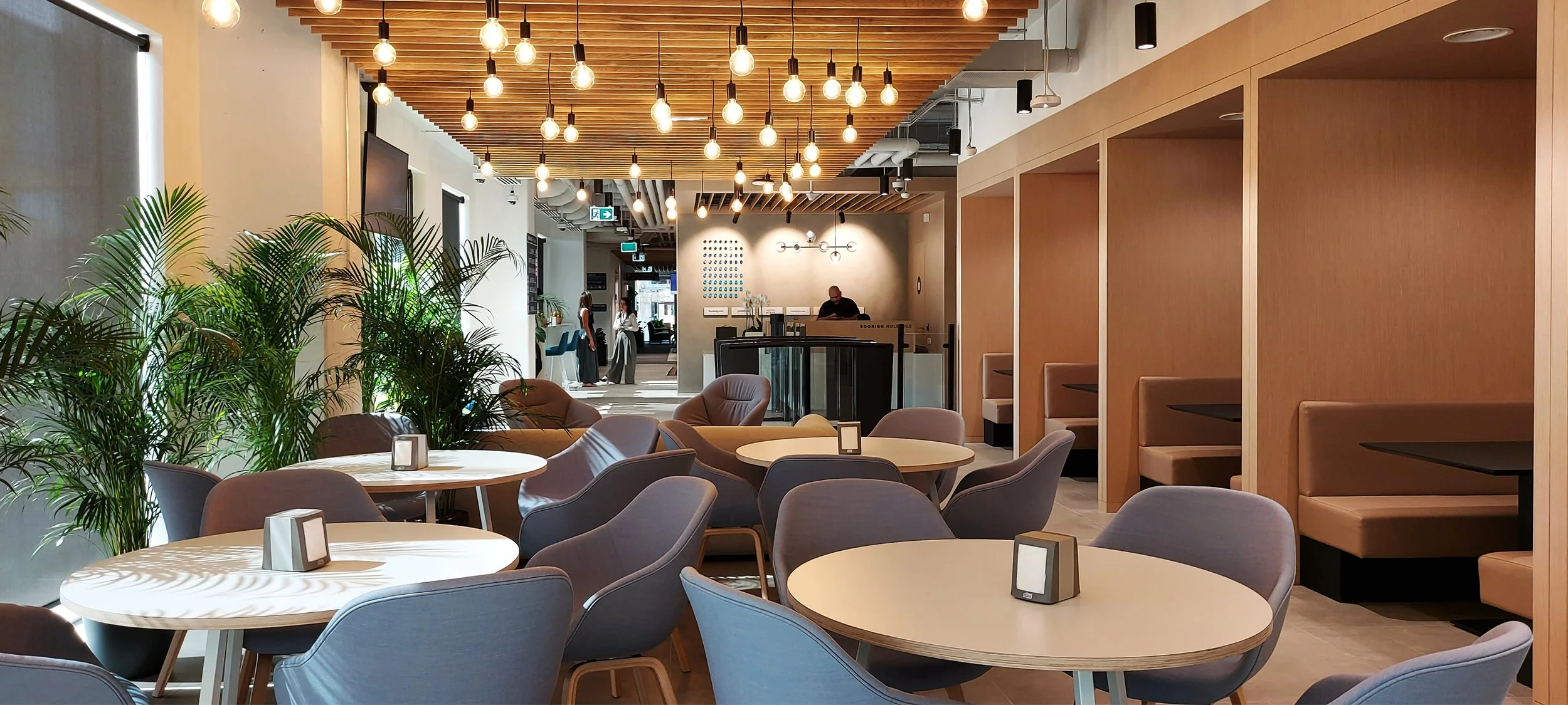 Modern cafe interior with round tables, plush chairs, green plants, and booth seating, illuminated by hanging Edison bulbs.