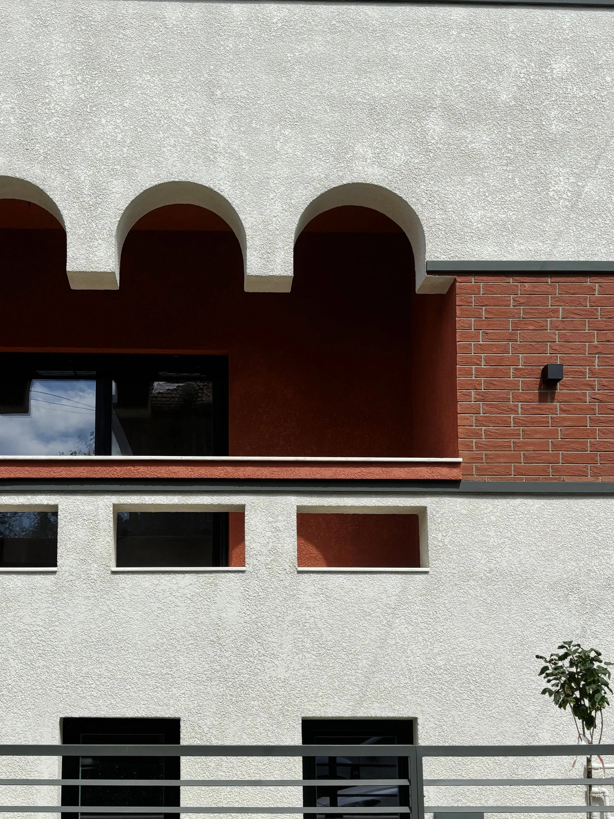 Close-up of a building facade featuring white textured stucco, red brick, and arched architectural details with a small tree in front.