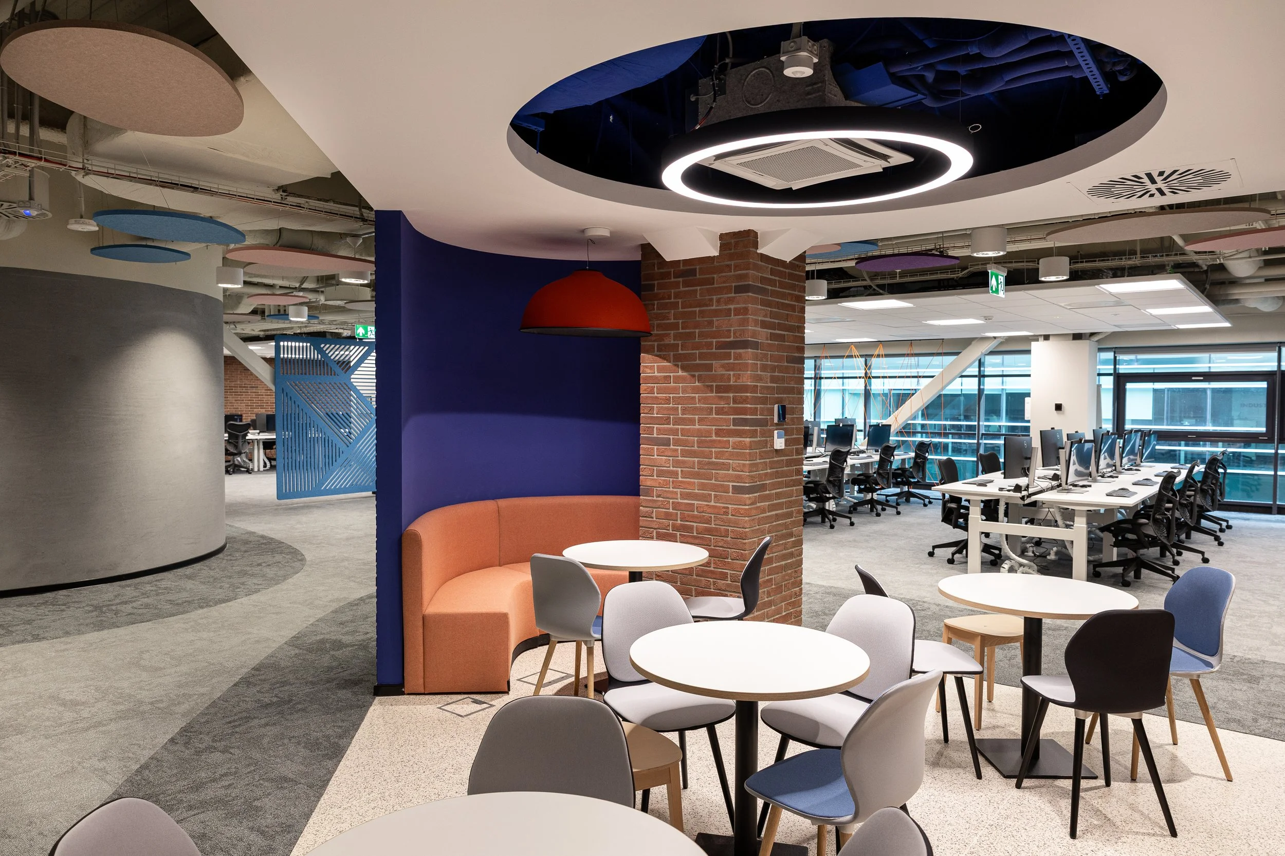 Modern office space with casual seating area featuring round tables and colorful chairs, open workstations with desktop computers, brick pillar, and large windows with city view.