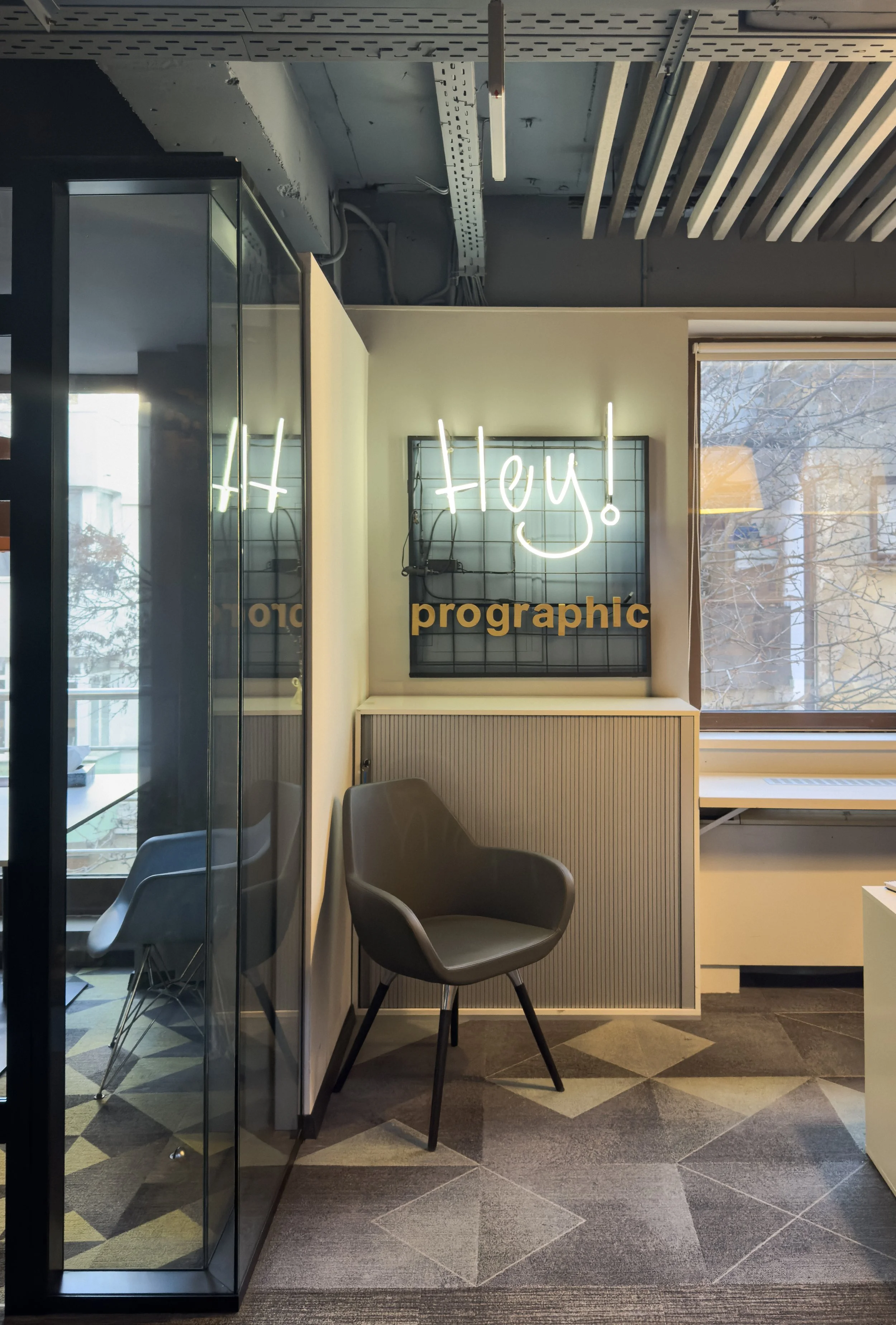 Modern office corner with a gray armchair, a window, and a neon sign on a grid wall that says 'Hey!' with part of the word 'graphic' visible below.