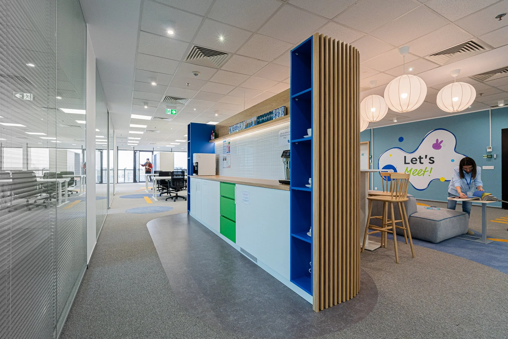 Modern office space with a reception desk, a lounge area, and large windows, decorated with a sign reading "Let's Meet!" on a blue wall.