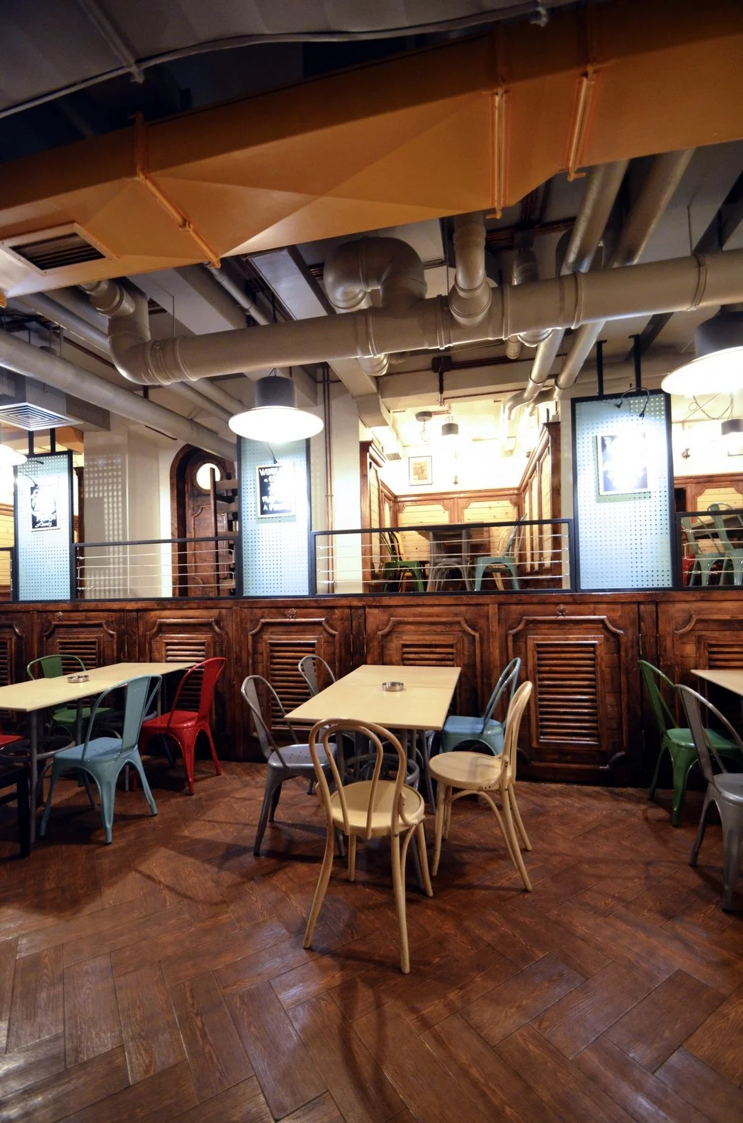 Interior of a cozy restaurant with wooden floors and colorful metal chairs around wooden tables, with exposed pipes and ducts on the ceiling and framed pictures on the walls.